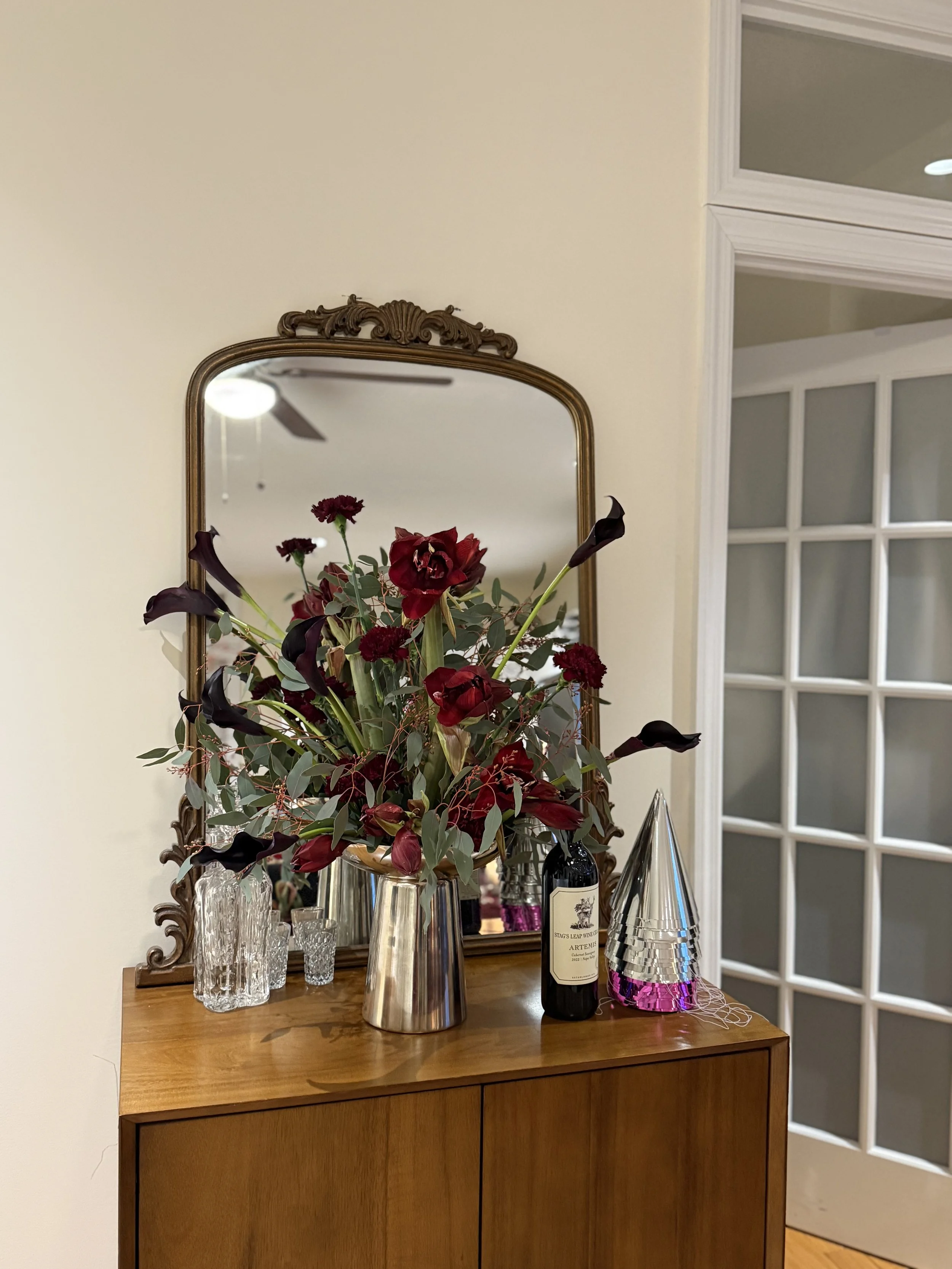 A wooden side table with a large floral arrangement, wine bottle, and party hats, reflected in a decorative mirror on a cream wall.