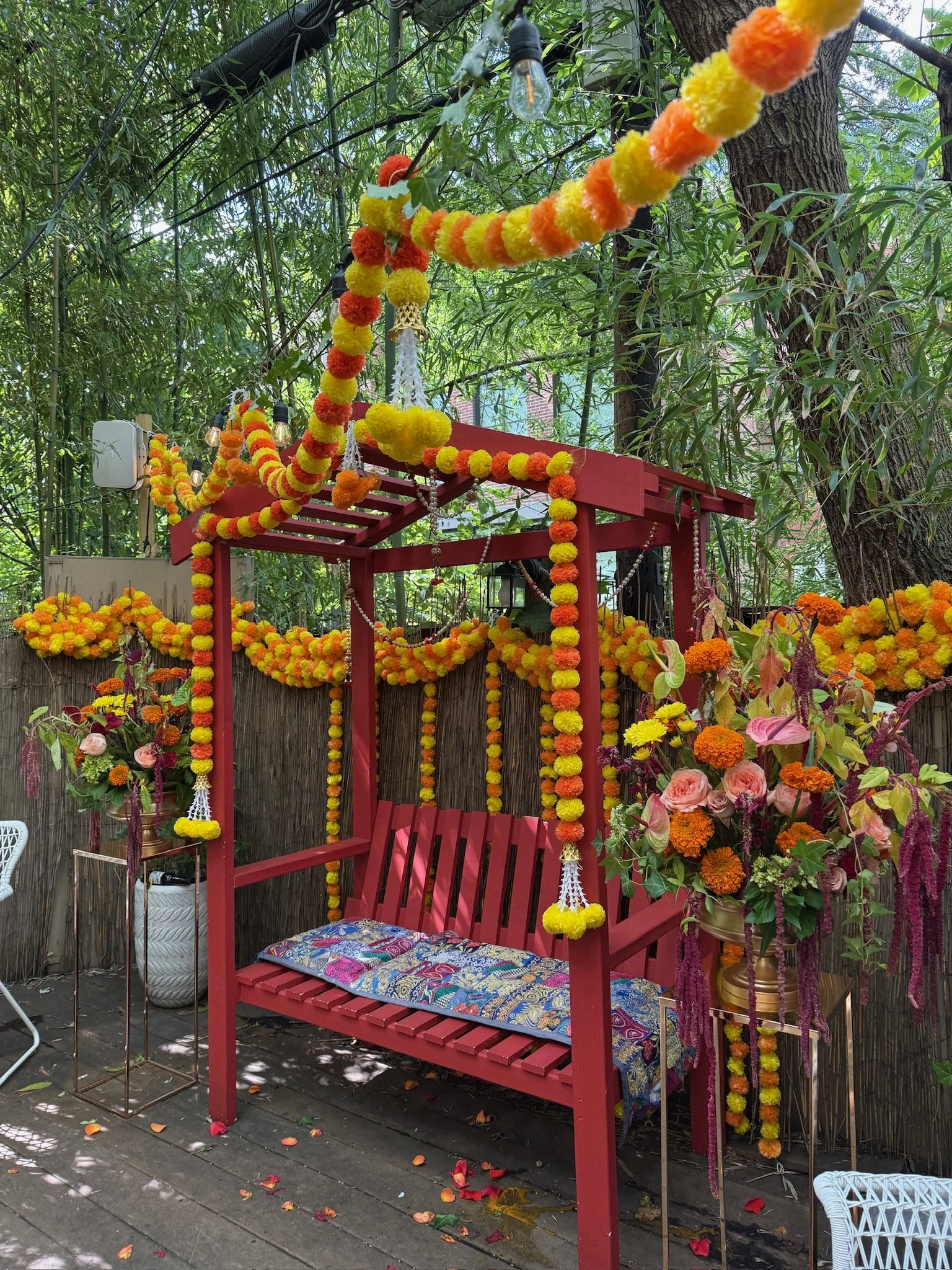 Decorated outdoor space with a red wooden bench, colorful flower garlands, hanging light bulbs, and an arrangement of flowers and plants on a wooden patio under trees.