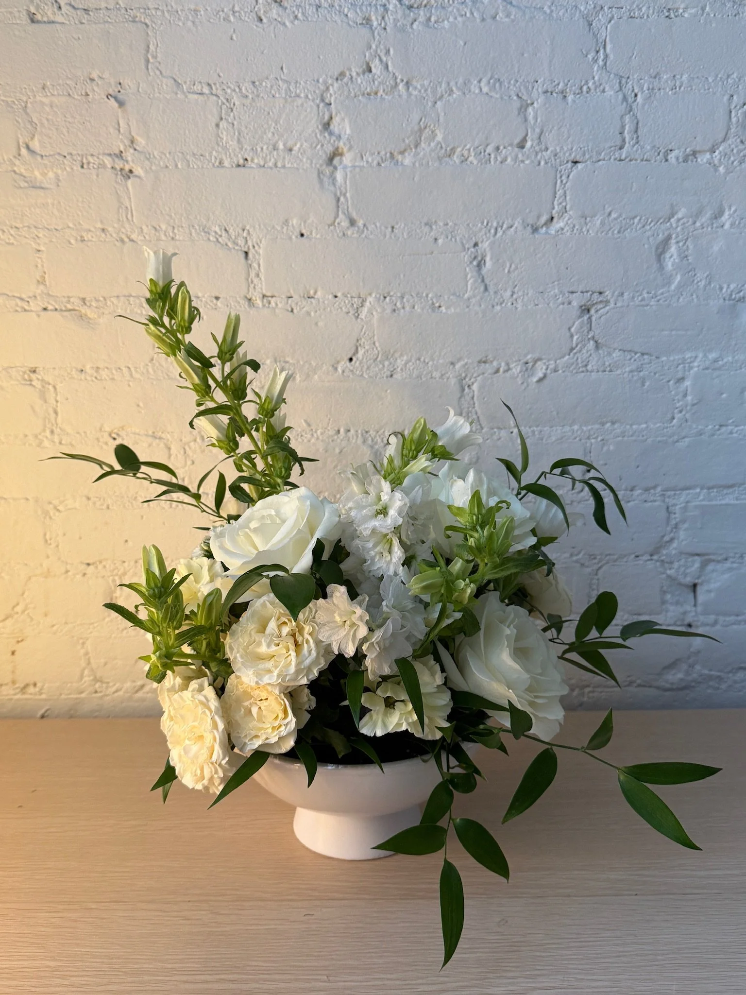 A floral arrangement with white roses, lilies, and greenery in a white vase against a white brick wall background.
