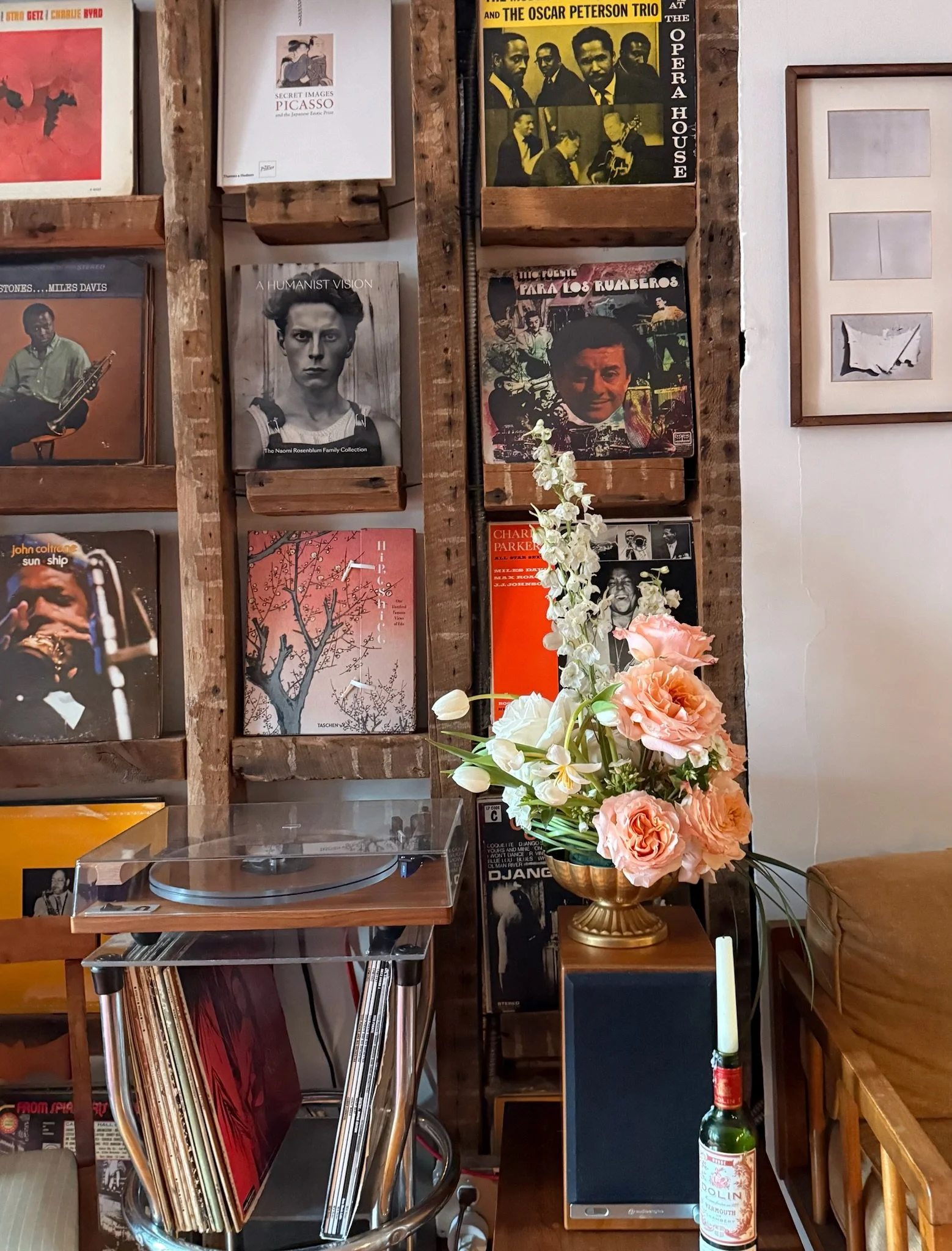 A room with a vinyl record collection on wooden shelves, a vintage turntable, a laptop, a large bouquet of pink and white flowers in a gold vase, and various posters and artwork on the wall, including a framed piece with three abstract sketches and p
