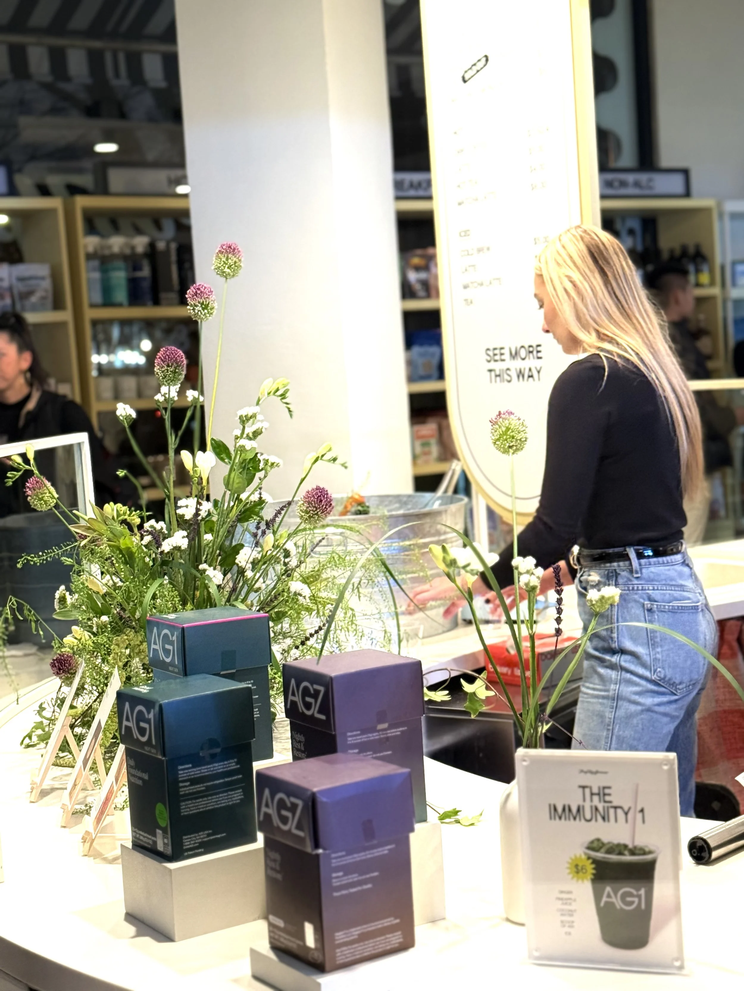 A woman with long blonde hair and black top at a counter with flowers and product boxes, in a retail space.