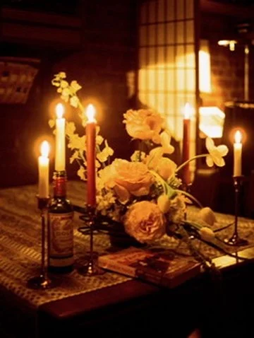A table with lit candles, a bouquet of yellow flowers, and a bottle, illuminated in warm light in a cozy room.