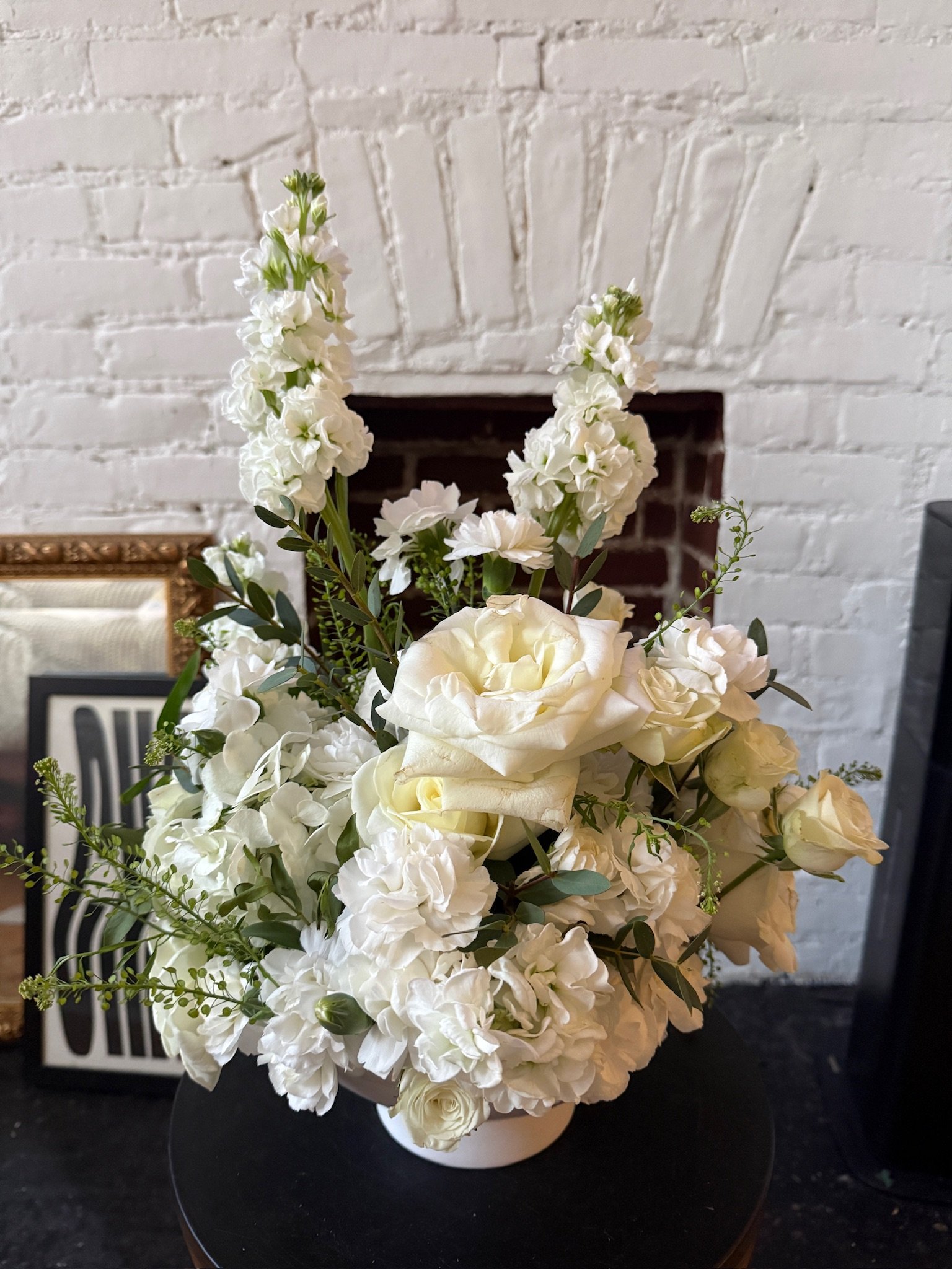 A bouquet of white flowers including roses and hydrangeas in a black vase on a black surface, with a white brick wall background.