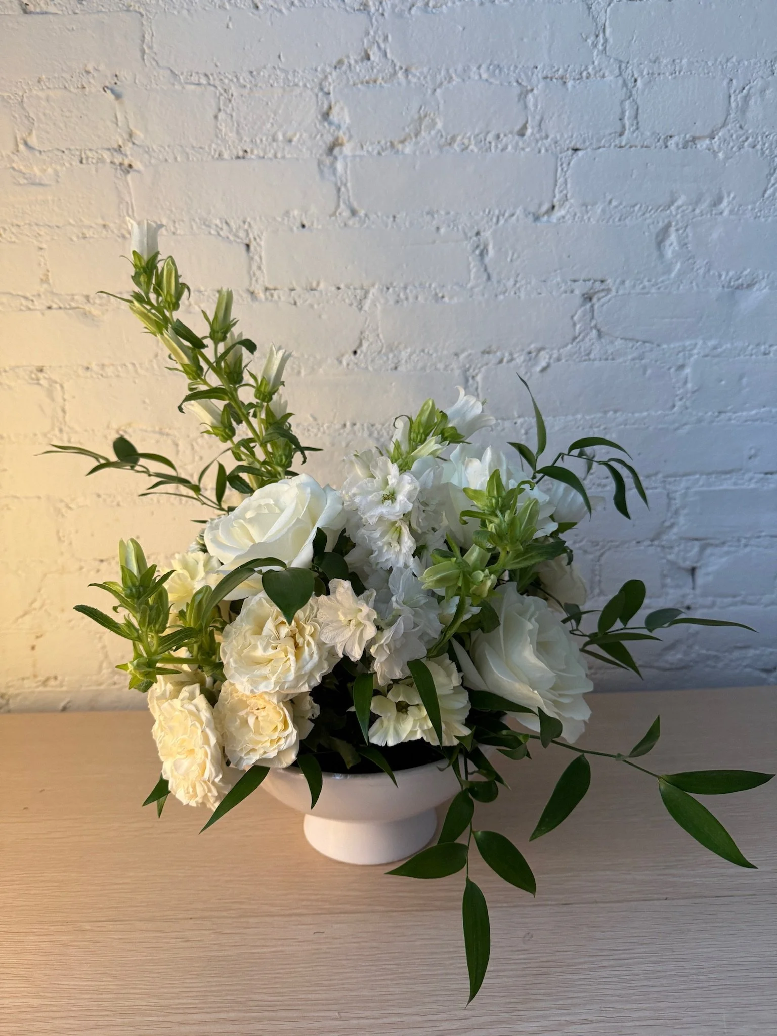 A white flower arrangement in a white vase placed on a light-colored wooden table against a white painted brick wall.