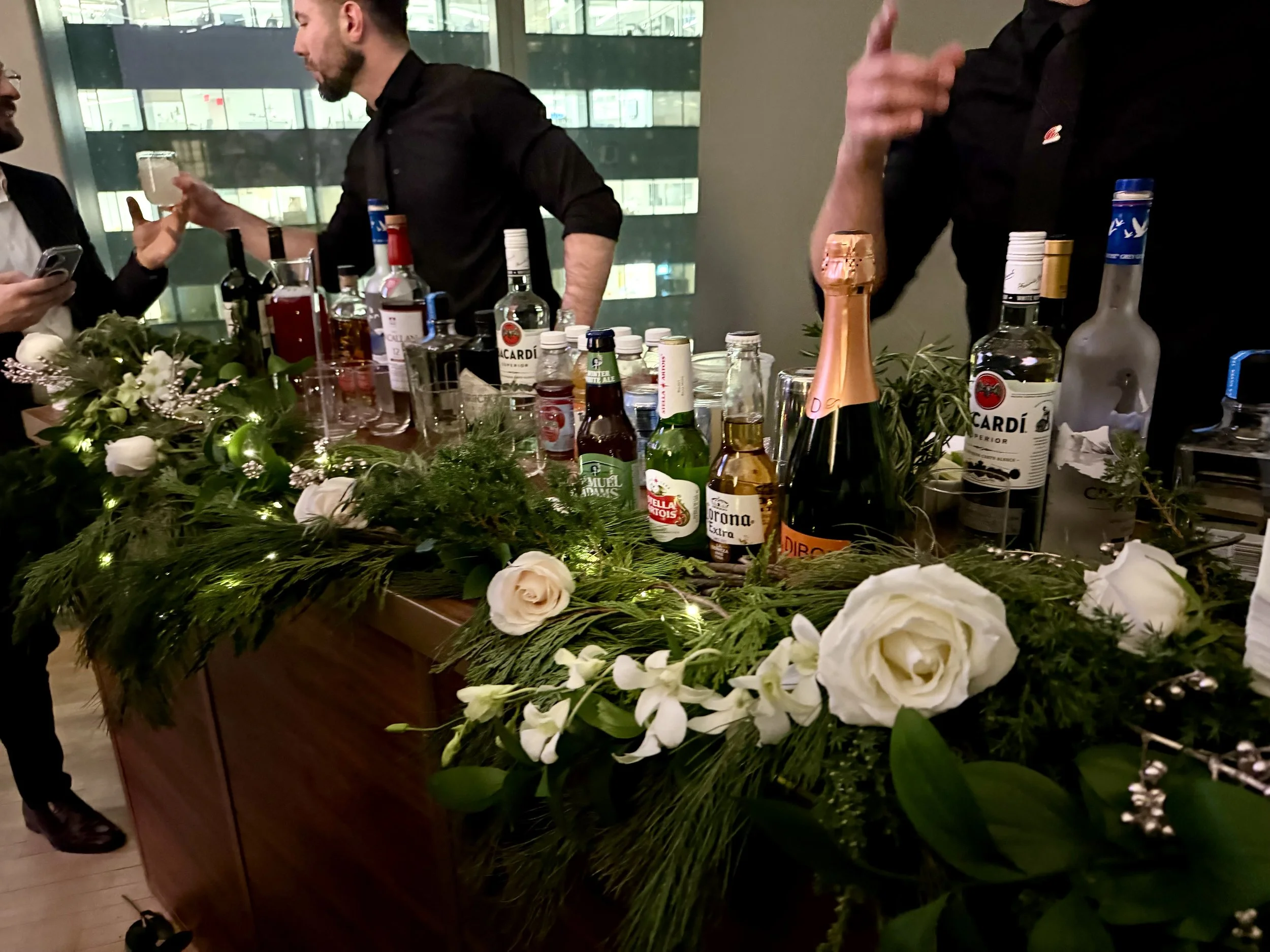 A bar decorated with green foliage and white flowers, with various bottles of alcohol, including Bacardi and Corona, and some people socializing in an indoor setting with city buildings visible through large windows in the background.