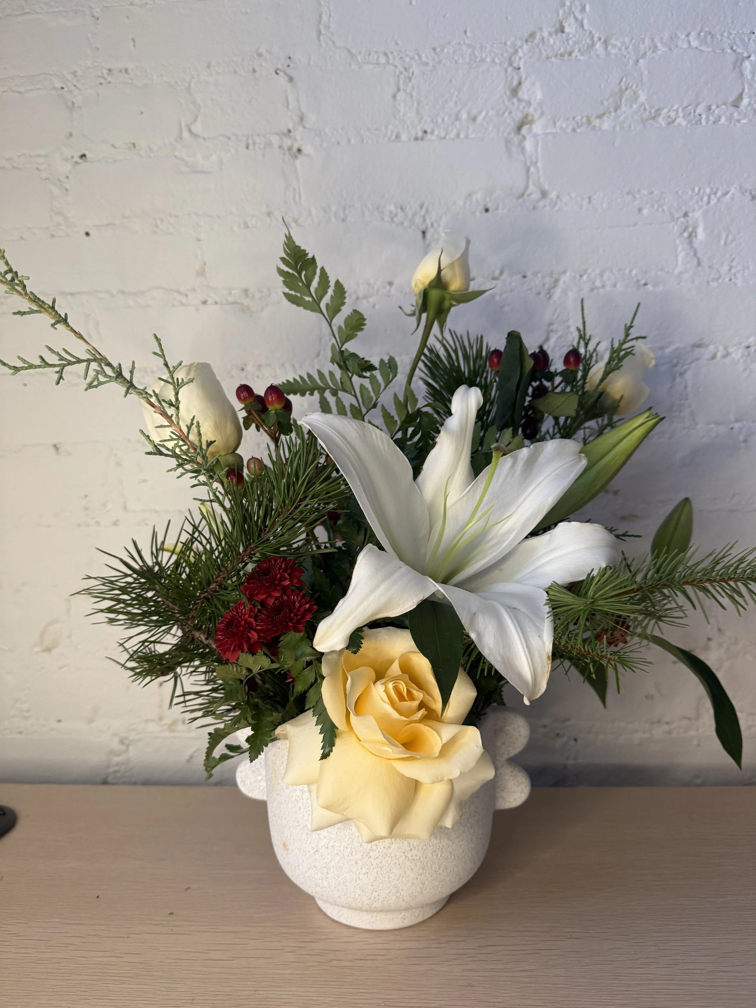 A floral arrangement with white lilies, yellow roses, red carnations, greenery, and small red berries in a white textured ceramic vase against a white brick wall.