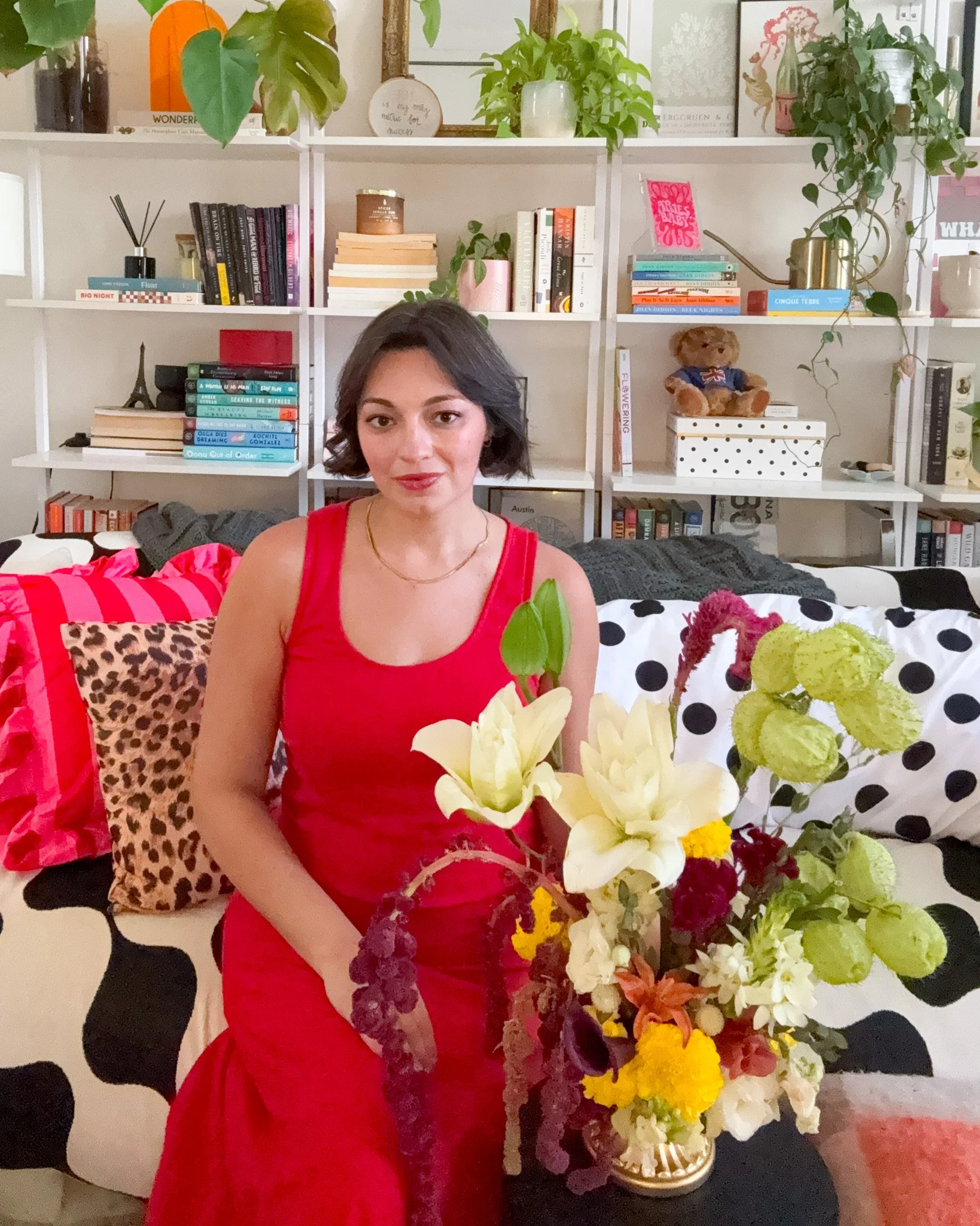 Woman with short dark hair and red dress sitting in front of a colorful flower arrangement in a cozy living room with bookshelves and decorative pillows.