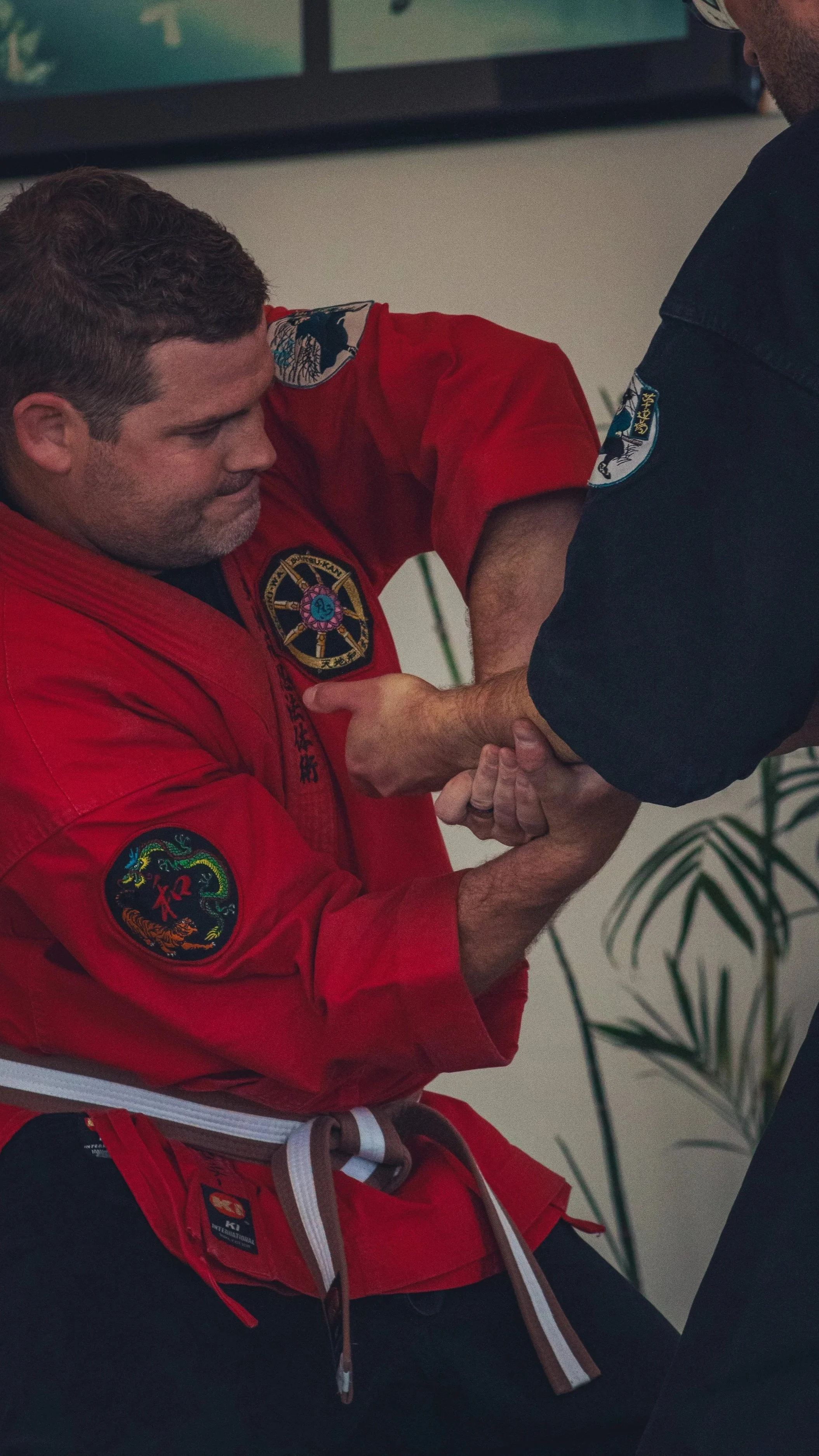 A person in a red martial arts gi with patches, including a dragon emblem, is practicing a martial arts technique with another person in a black gi. The person in red is gripping the other's wrist while bending the arm.