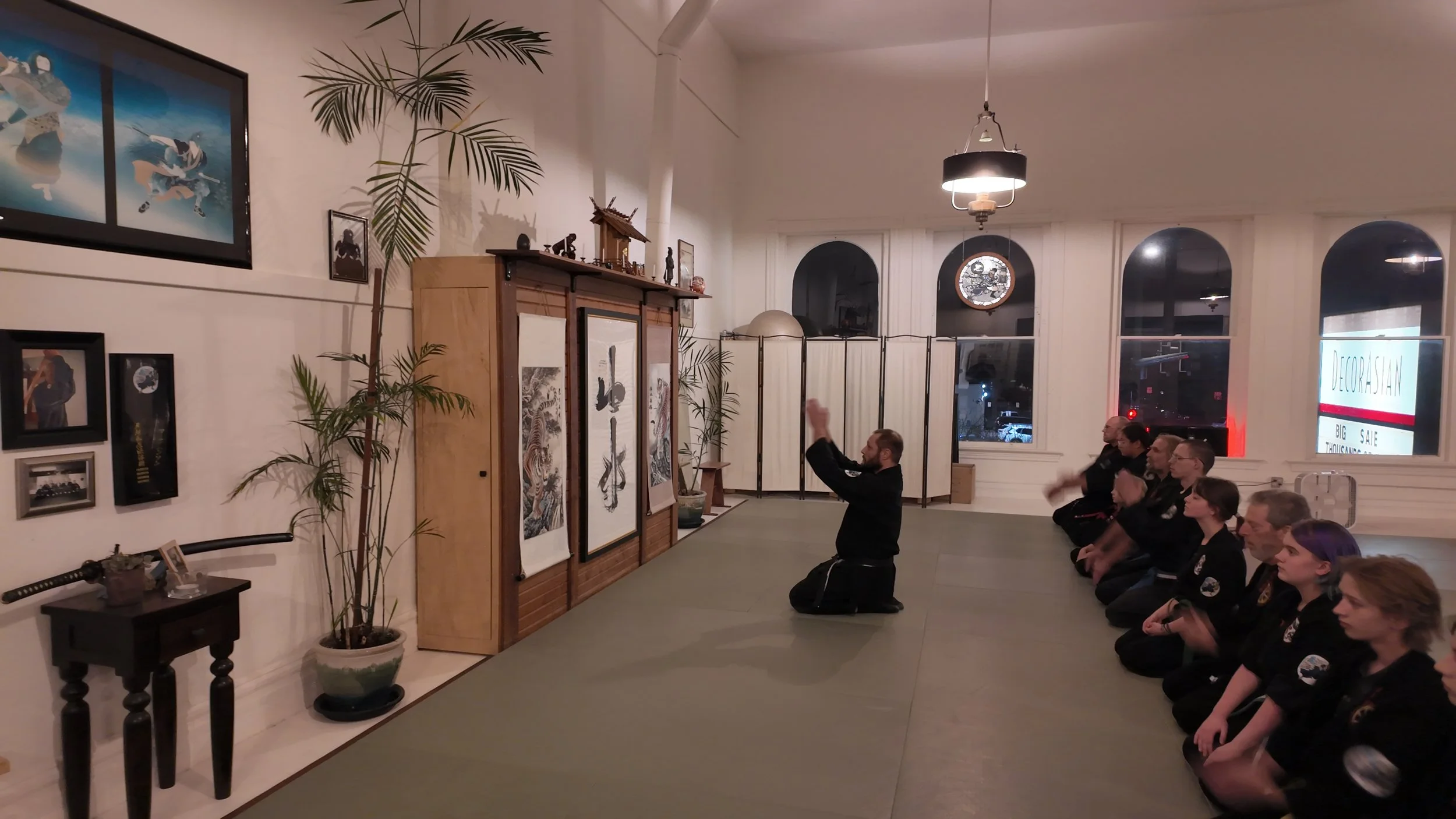 A group of martial arts students in black uniforms and a kneeling instructor in black uniform practicing karate or judo in a studio decorated with Japanese artwork, plants, and framed photos, during an evening class.