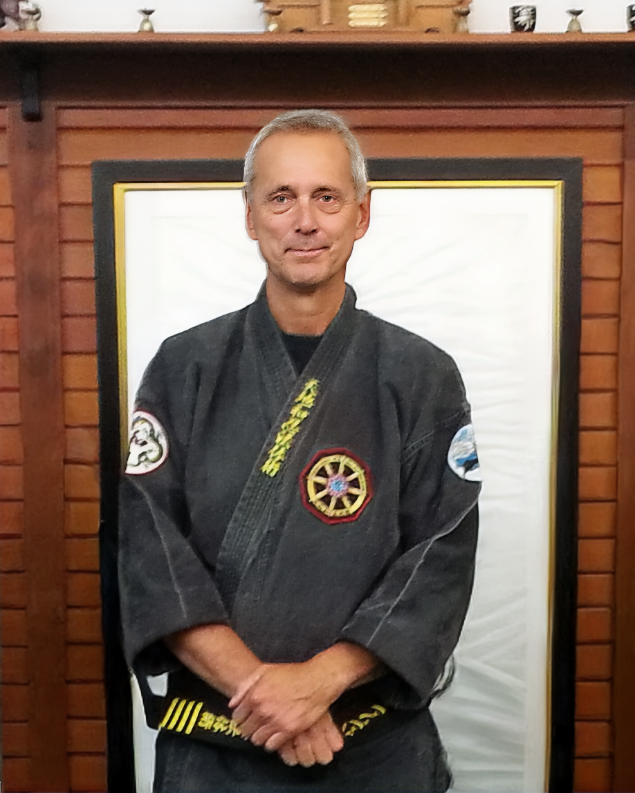 Sensei Marc, a man in a black martial arts uniform standing in front of a wooden wall and white background, with patches on his uniform indicating martial arts rank and affiliation.