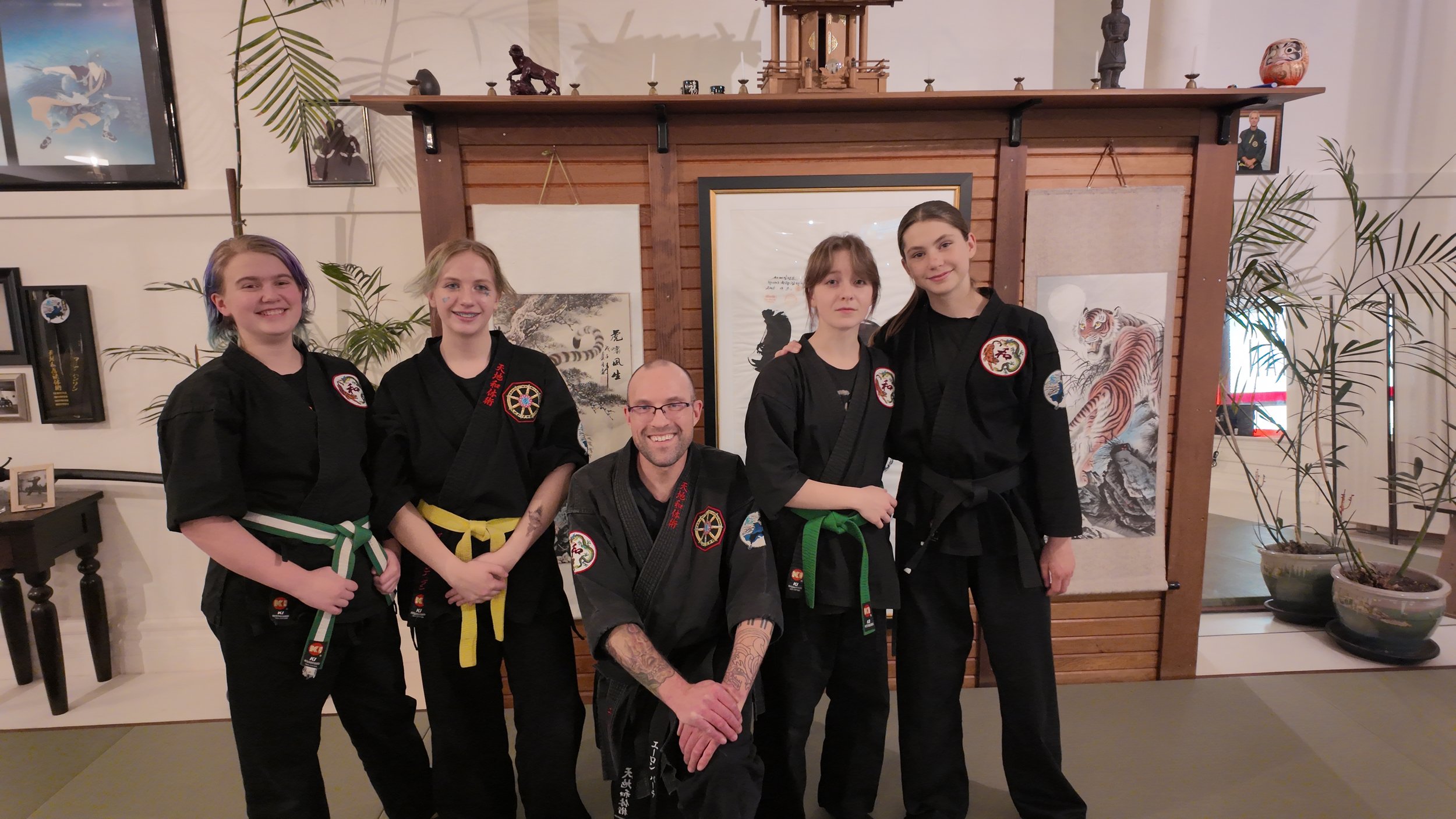 Group of five people in martial arts uniforms posing indoors, with a wooden partition and traditional artwork in the background.