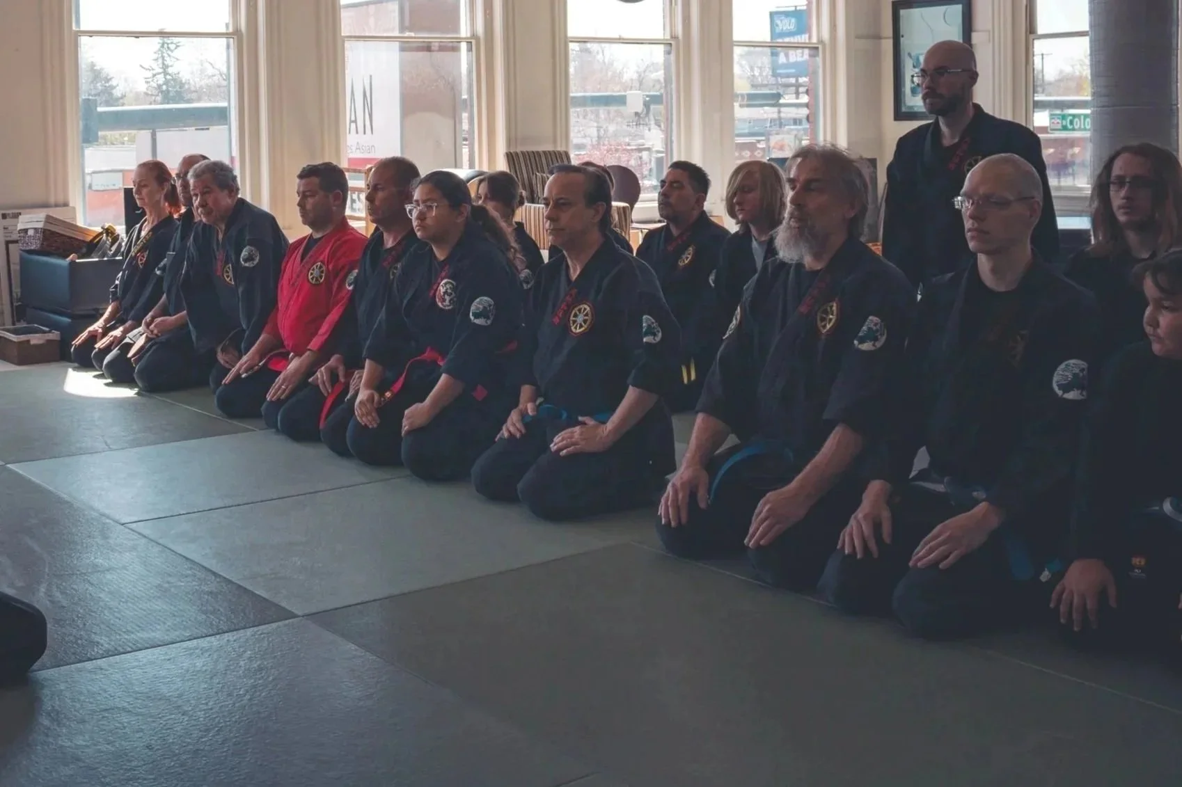 A group of martial artists kneeling on mats inside a dojo, dressed in black uniforms with colorful patches and belts, with sunlight coming through large windows behind them.