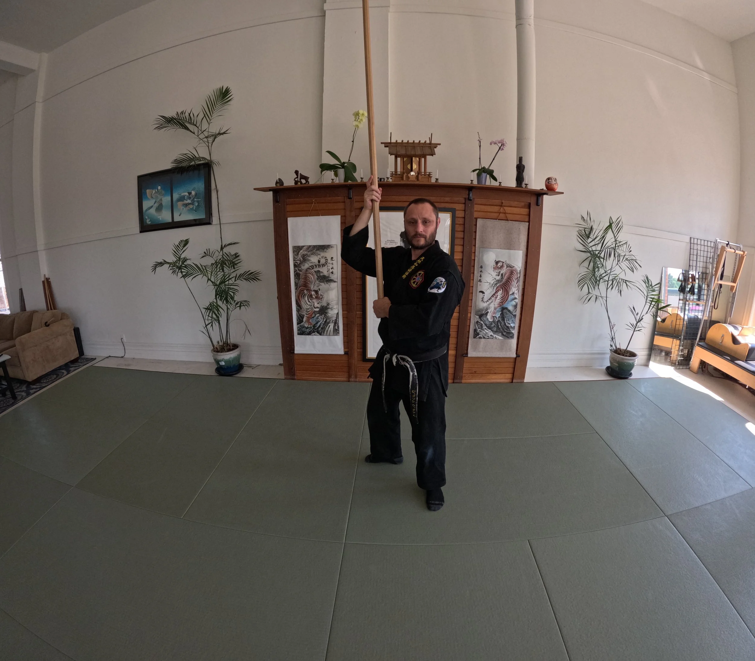 Man dressed in martial arts gi holding a long wooden staff inside a martial arts dojo with artwork and plants in the background.