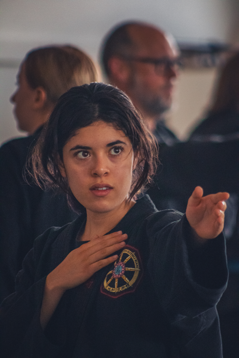 Young woman with dark hair in a ponytail, wearing a black martial arts uniform with a patch, extending her right arm forward in a martial arts stance, with a serious expression. Two blurred background adults, one with glasses, visible behind her.