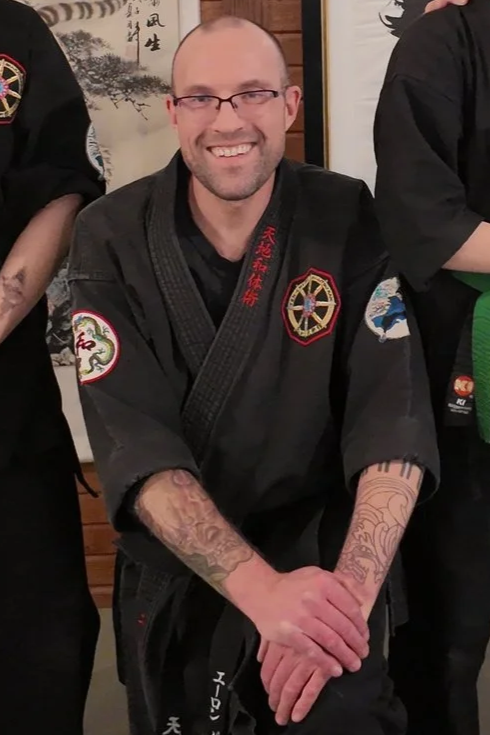 Man smiling, wearing a black martial arts uniform with patches, sitting with hands resting on his knees, surrounded by others also in martial arts uniforms.