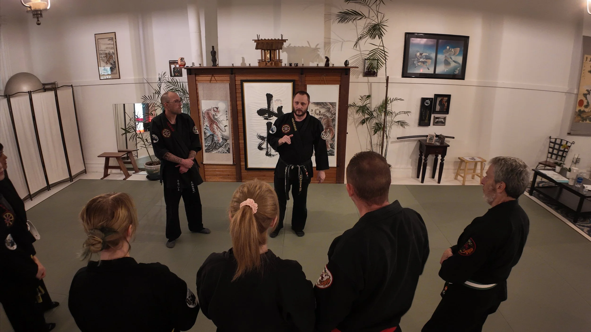Martial arts instructor teaching students in a dojo with traditional Japanese artwork and scrolls on the wall.