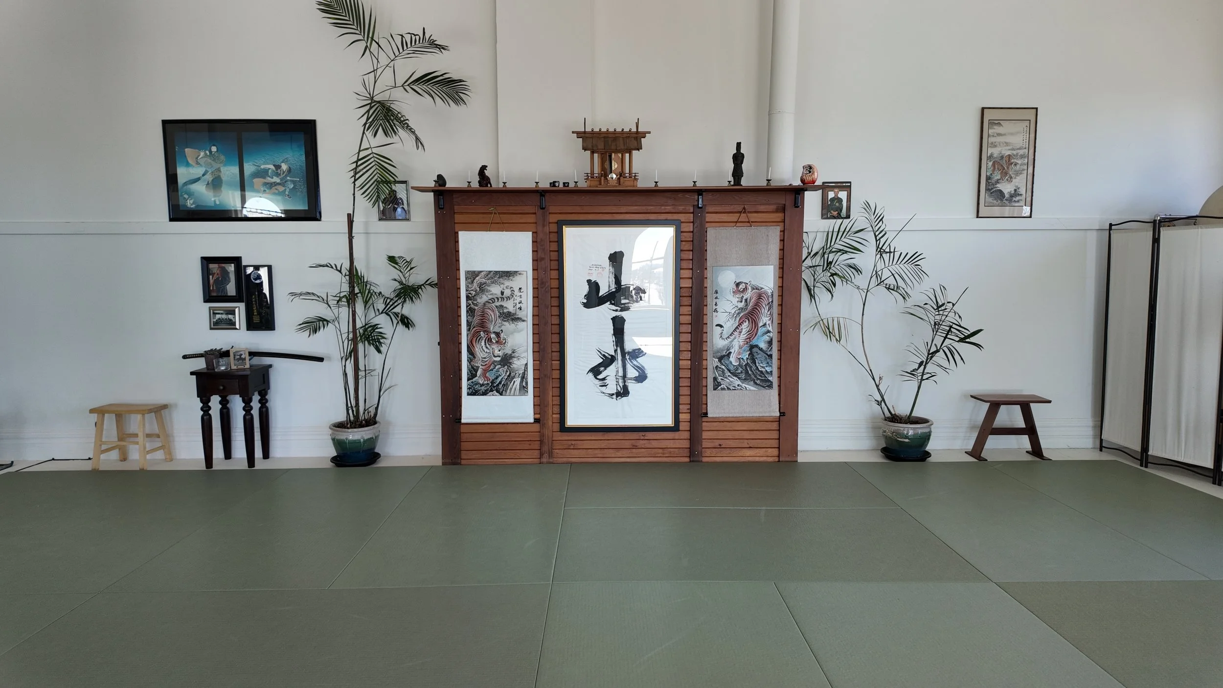 Interior of a Japanese-style room with a wooden display case, hanging scrolls, framed artwork, potted plants, and small tables.