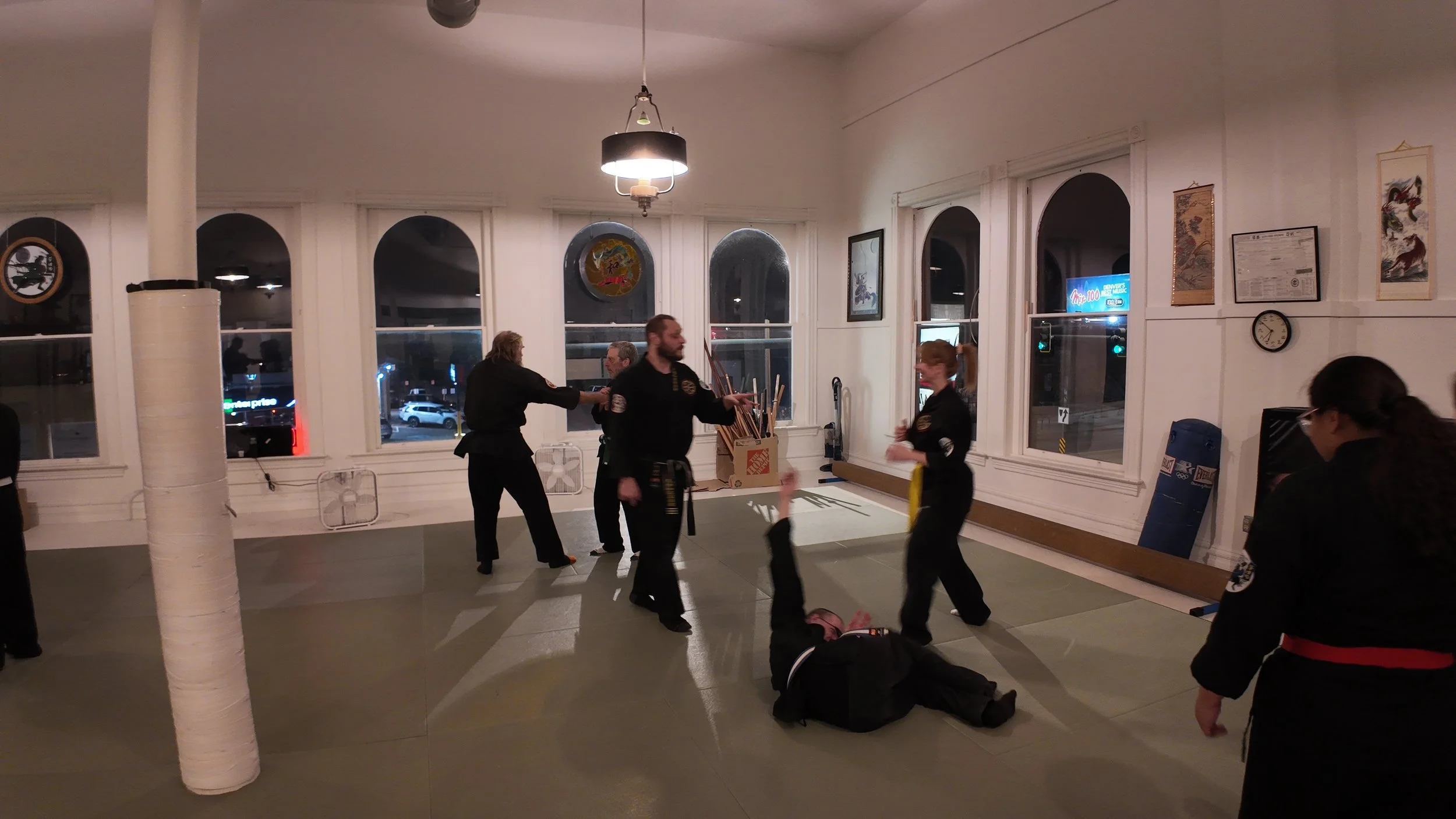Martial arts class with students practicing fighting techniques on mats in a well-lit room, with large windows, framed pictures, and a clock on the wall.