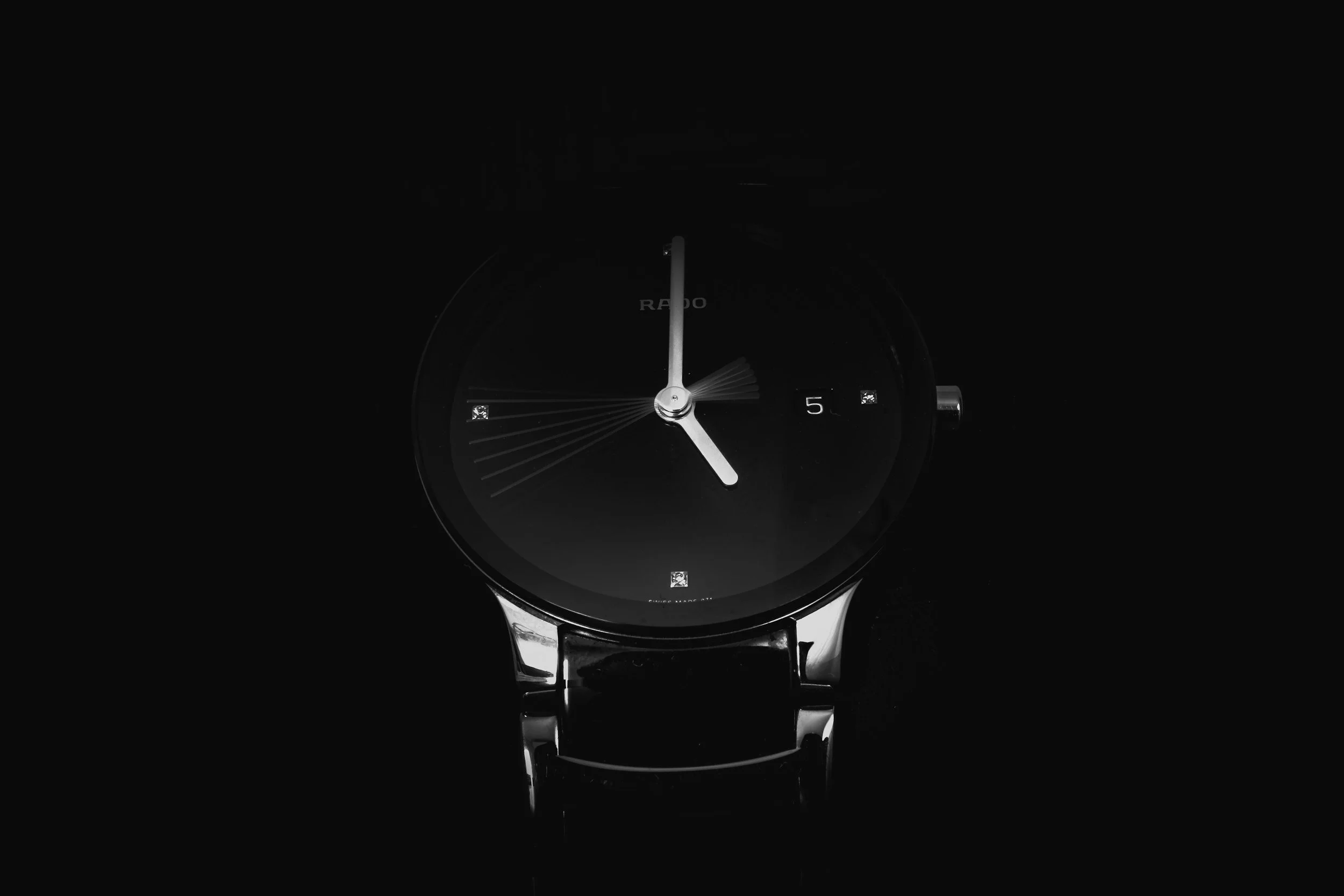 A black and silver wristwatch with a black face, silver hour and minute hands, and a date window showing the number 5, against a dark background.