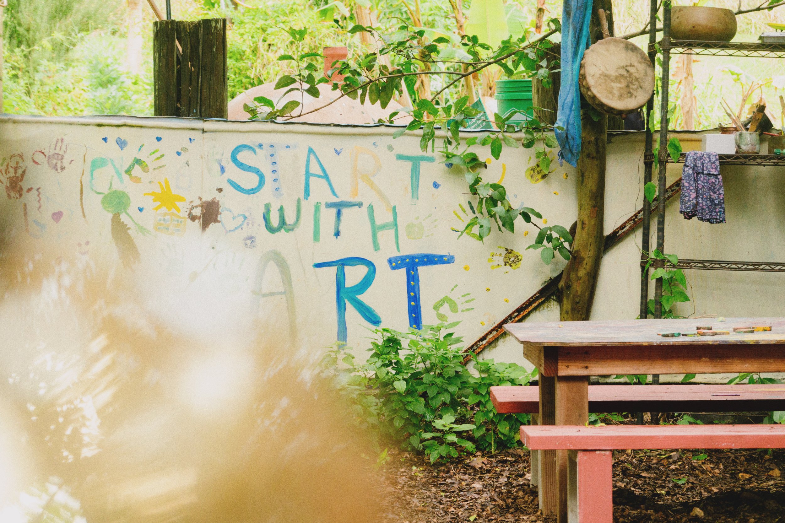 This image shows an outdoor area with a painted white wall that has colorful drawings and the words 'SART WITH ART' on it. There are wooden benches with red seats and a metal shelving unit with various items. Green foliage and trees are visible in the background, suggesting a garden or natural setting.