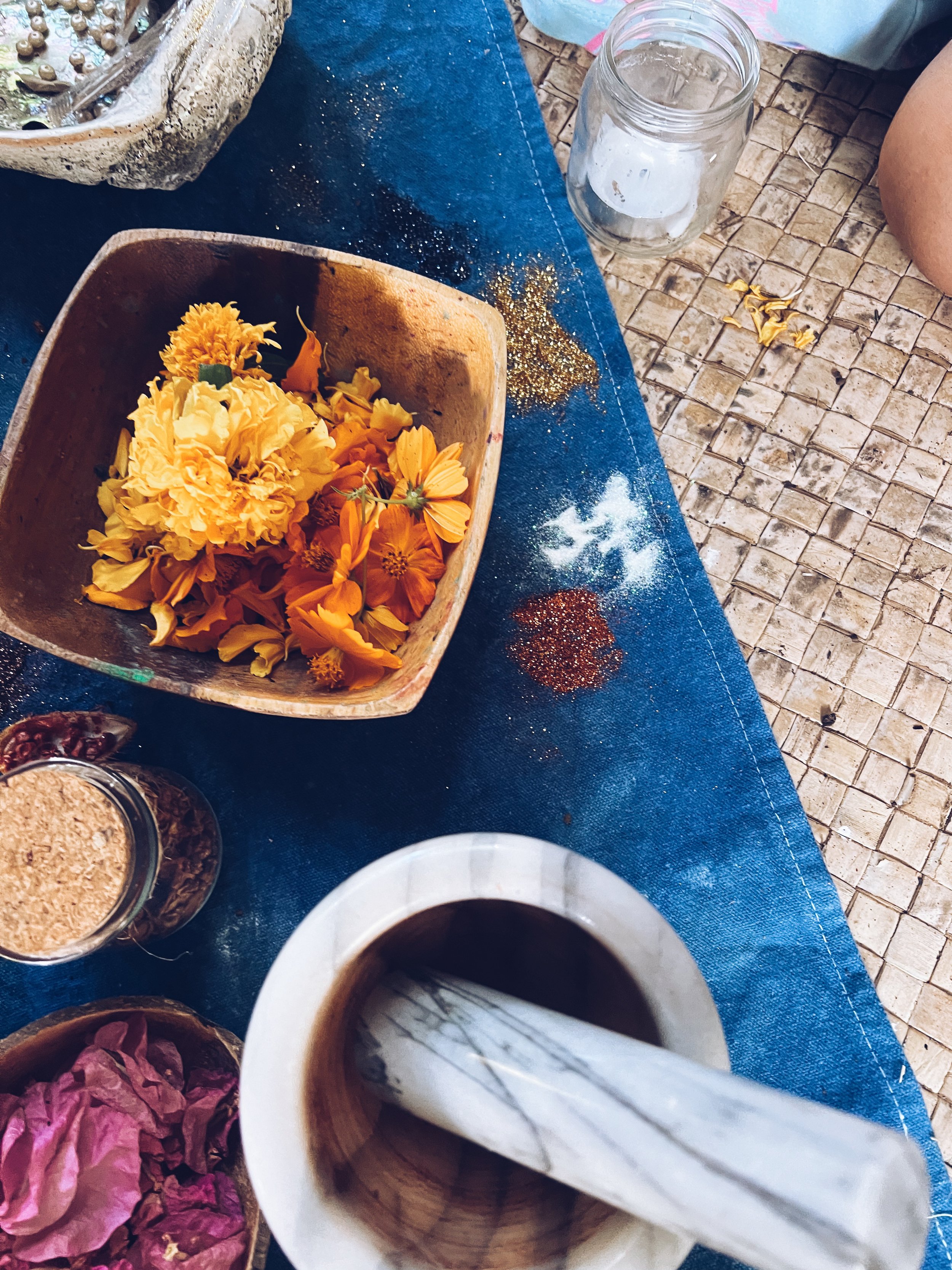 A table with yellow and orange flowers in a wooden bowl, spices, a glass jar with ice, a cup of coffee, and a brown paper bag, with part of a person's arm visible.