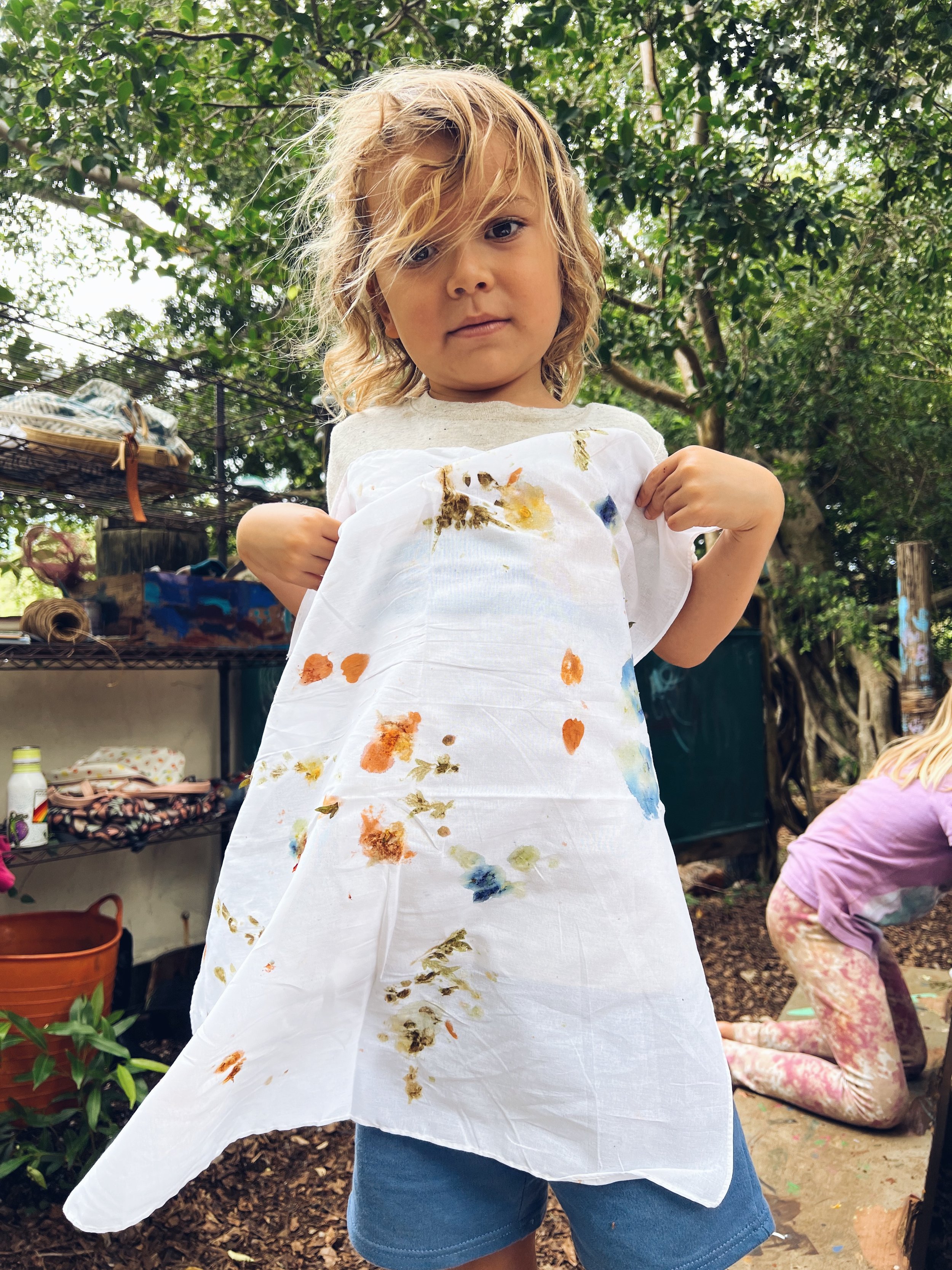 A young girl with curly blonde hair wears a white apron covered in colorful paint stains. She is outdoors amidst green trees, with her arms raised and hands gripping the apron to show off the paint. There is another child sitting on the ground to her