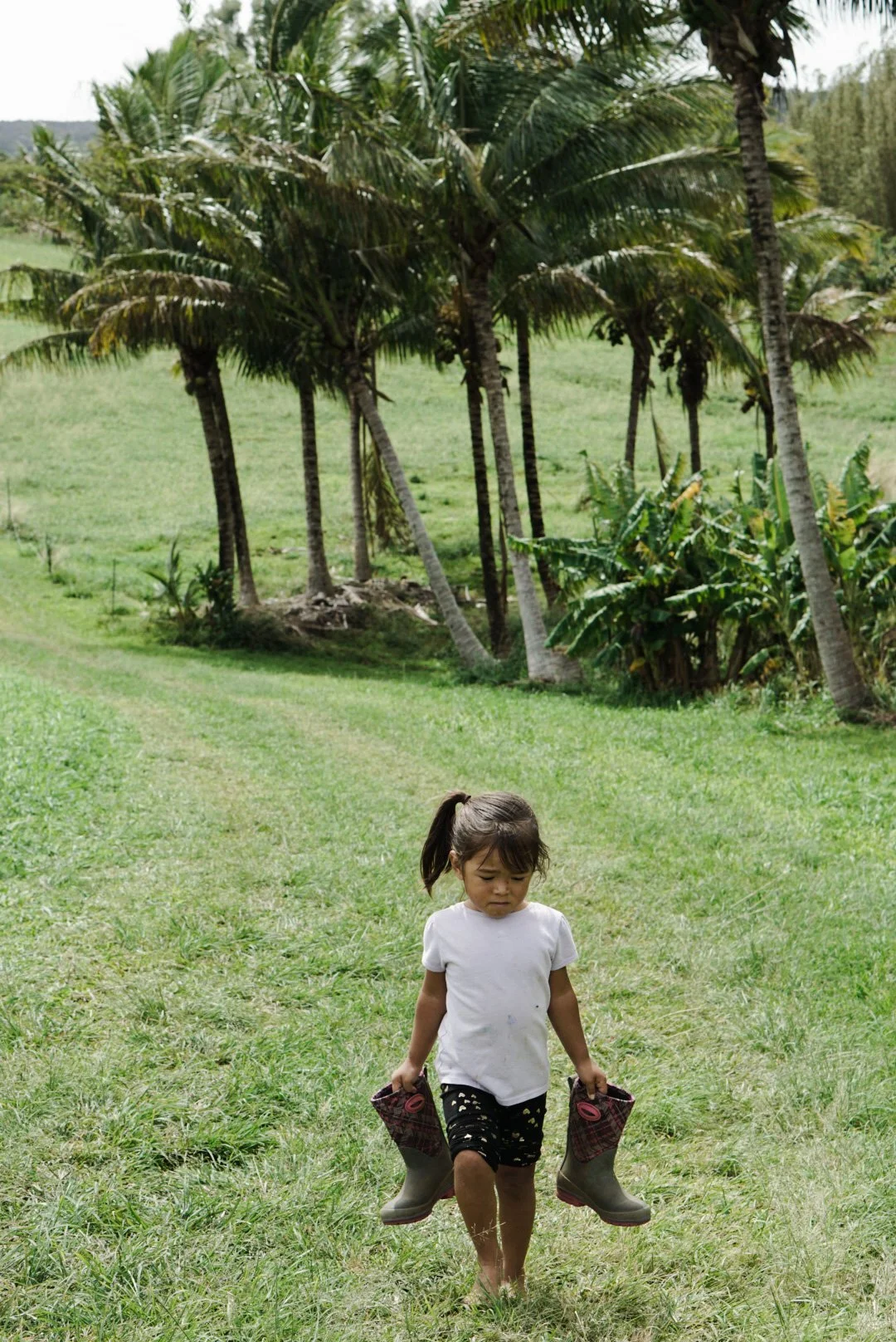 A young girl in a white shirt and black shorts walking on grass while carrying a pair of rain boots in each hand in a lush green landscape with palm trees.