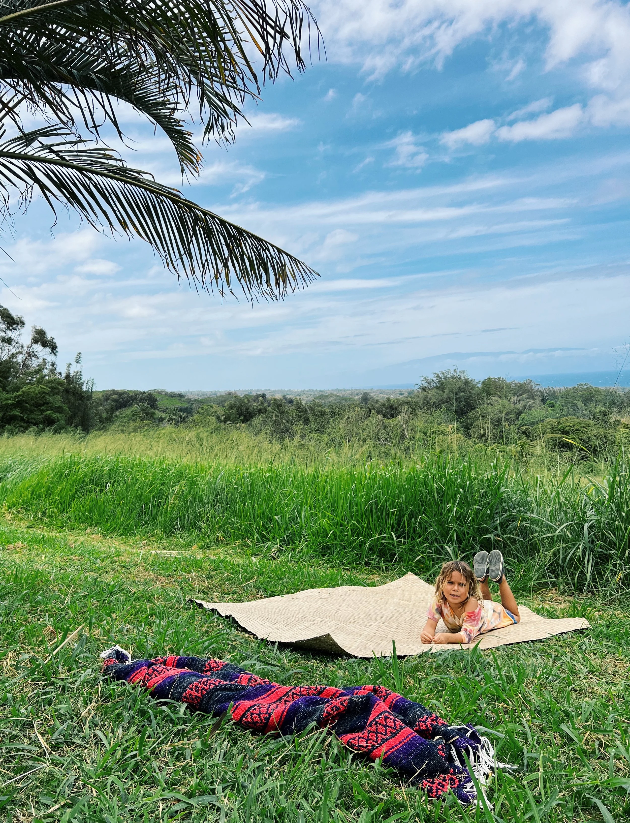A young girl with curly blonde hair lying on her stomach on a beige mat on green grass, with her feet raised and wearing colorful clothing, in a scenic outdoor landscape with lush greenery, trees, and a blue sky with clouds.