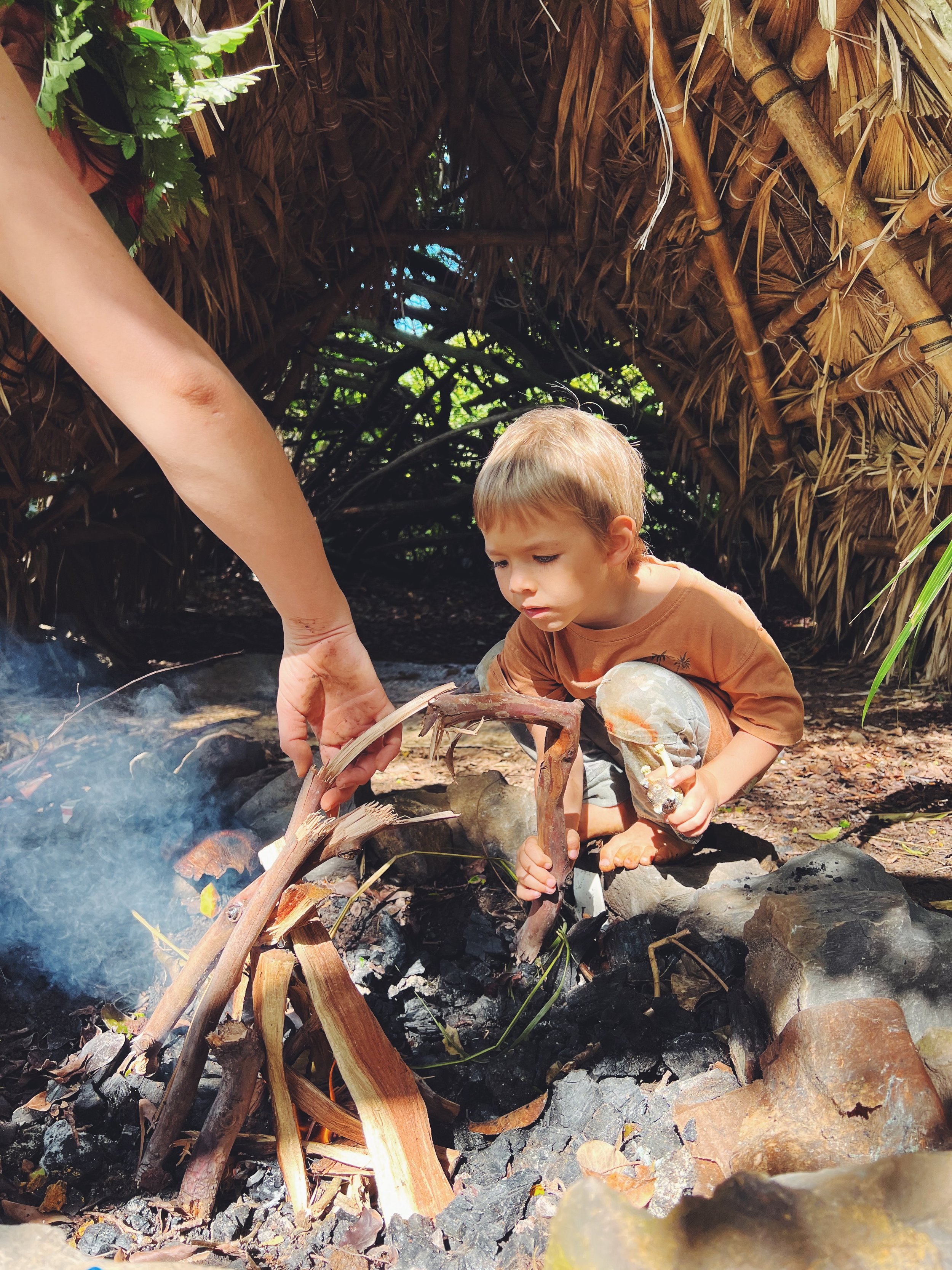 A young child squats near a small campfire made of rocks and wood inside a thatched shelter, surrounded by greenery.