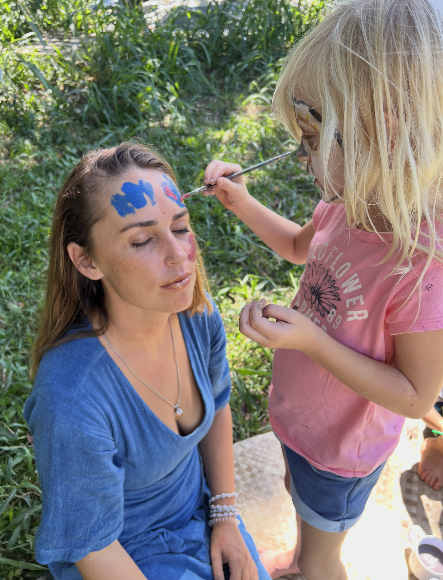 A young girl with face paint is painting a woman’s face with blue and red colors outside in a grassy area. The woman has her eyes closed and is wearing a blue top, while the girl is in a pink shirt and shorts, with her face also painted.