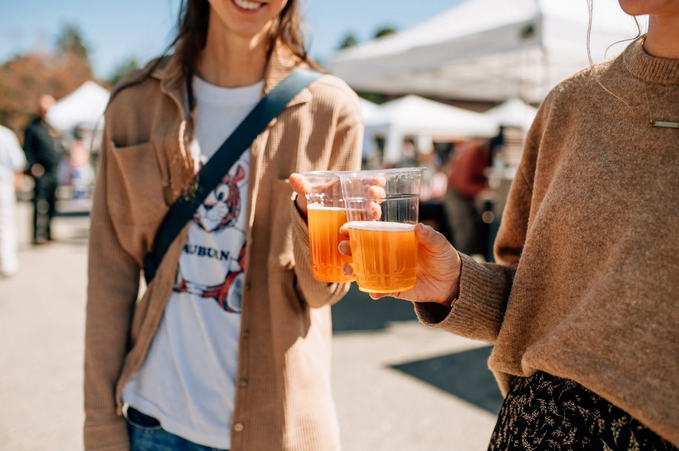 Two women holding plastic cups of orange beer at an outdoor event or festival, with tents and other people in the background.