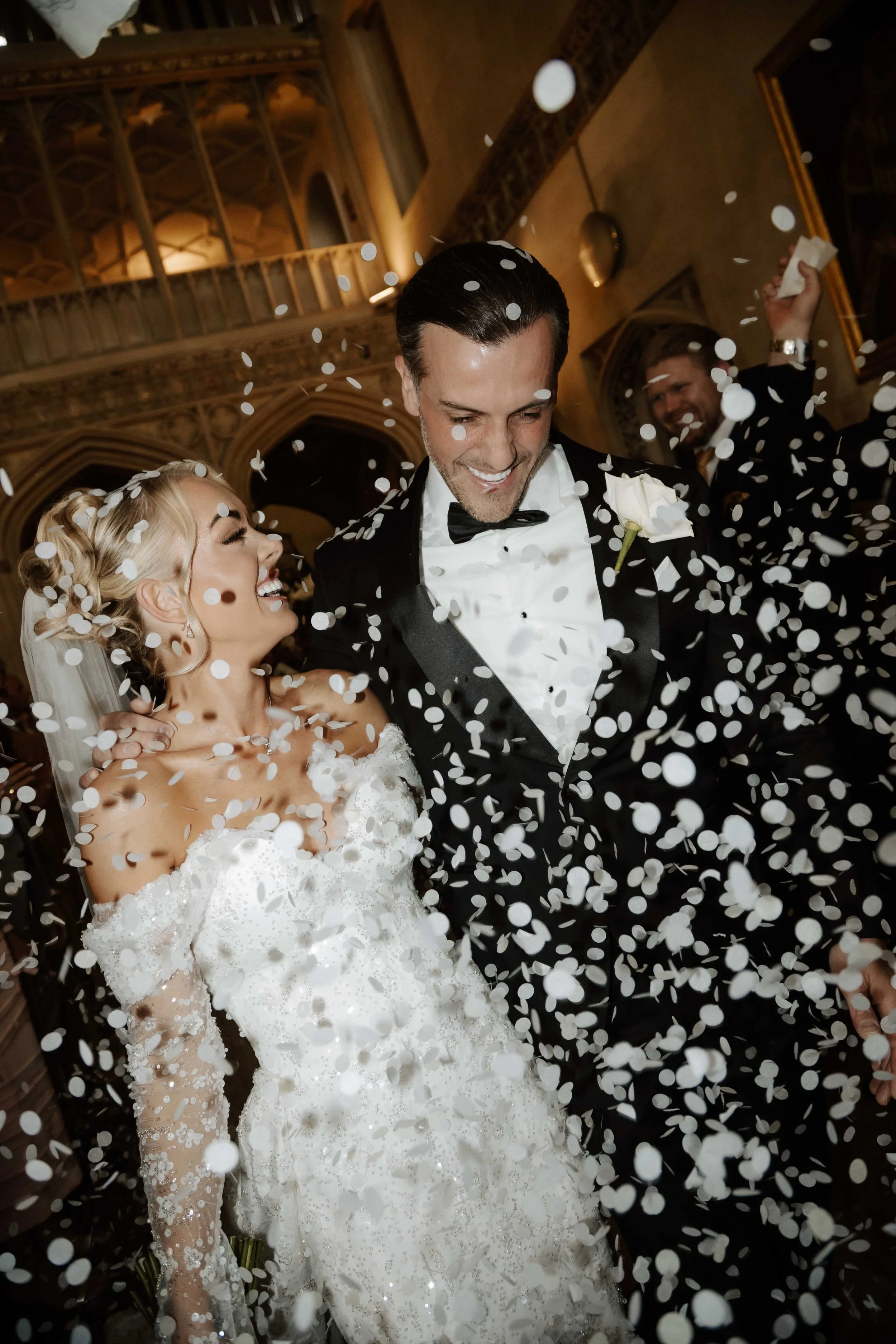 A newlywed couple celebrating their wedding with white confetti falling around them. The bride is wearing a white lace wedding gown and the groom is in a black tuxedo with a white boutonniere. They are smiling joyfully inside an ornate, elegant venue.