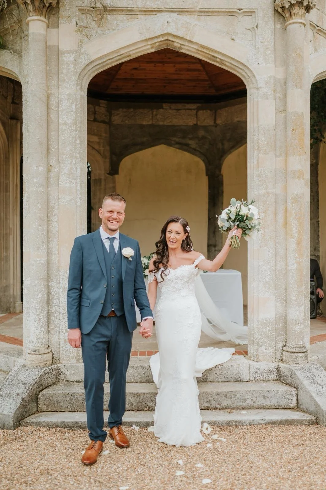 A newlywed couple holding hands and smiling, standing under a stone archway of a historic building; the bride is holding a bouquet of white flowers, wearing a white wedding dress, and the groom is in a blue suit and brown shoes.