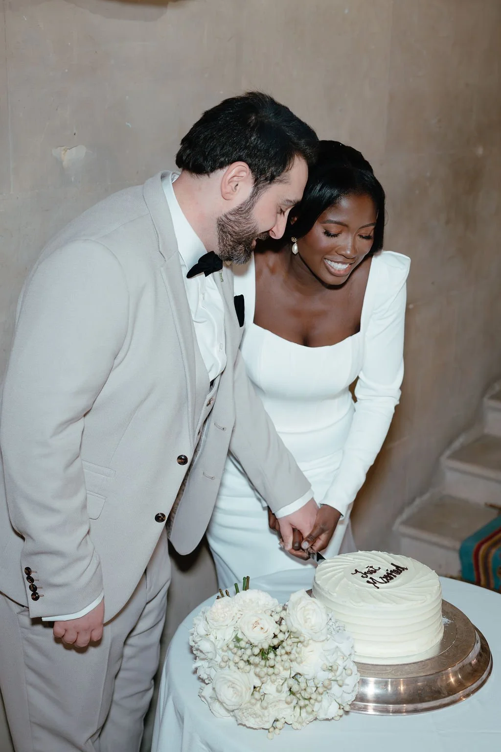 A newlywed couple, dressed in wedding attire, cutting their wedding cake together. The groom has a beard and is wearing a light gray suit with a black bow tie, and the bride has dark skin and black hair, wearing a white dress with puffed sleeves.