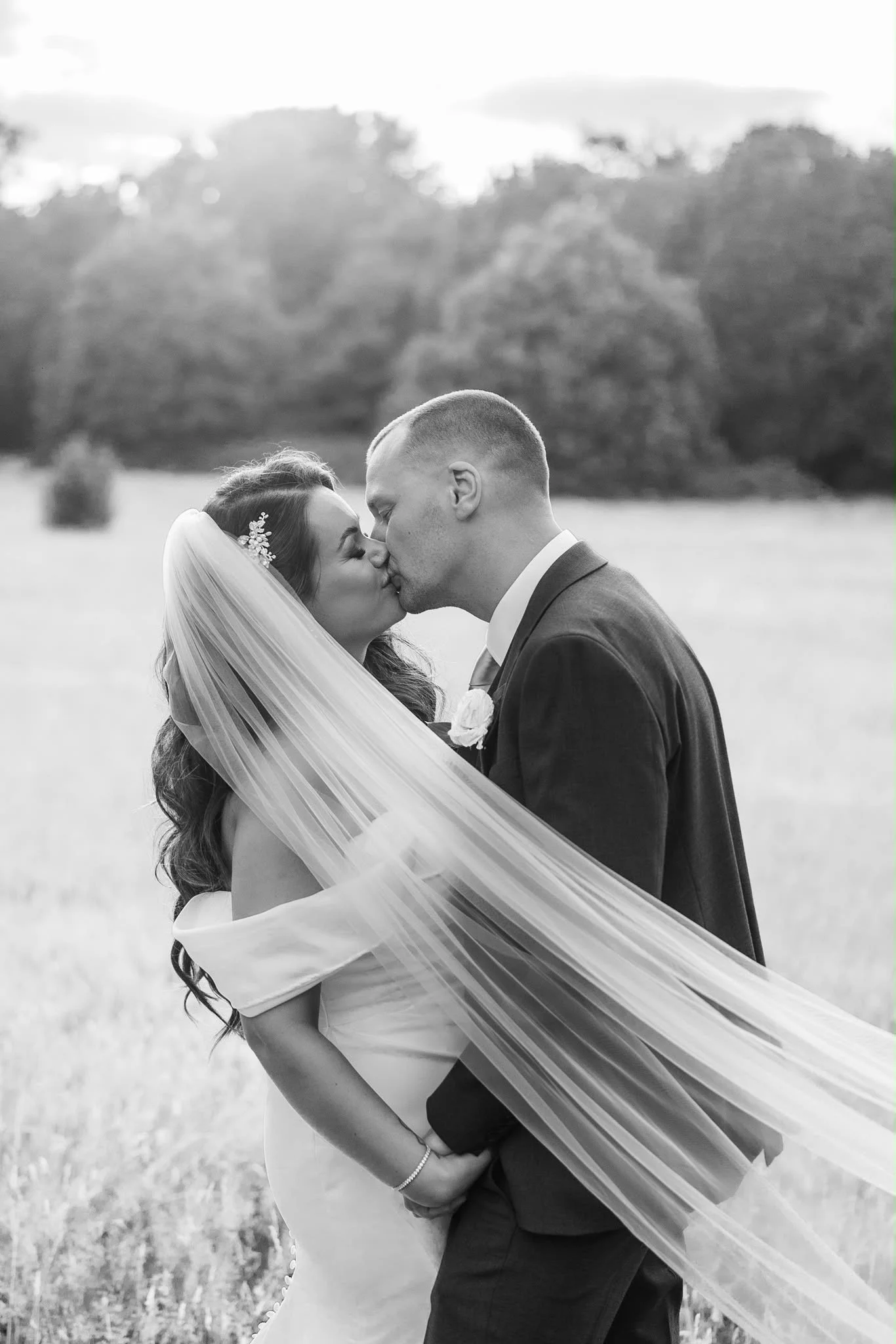 A black-and-white photo of a bride and groom kissing outdoors, with trees and a cloudy sky in the background.