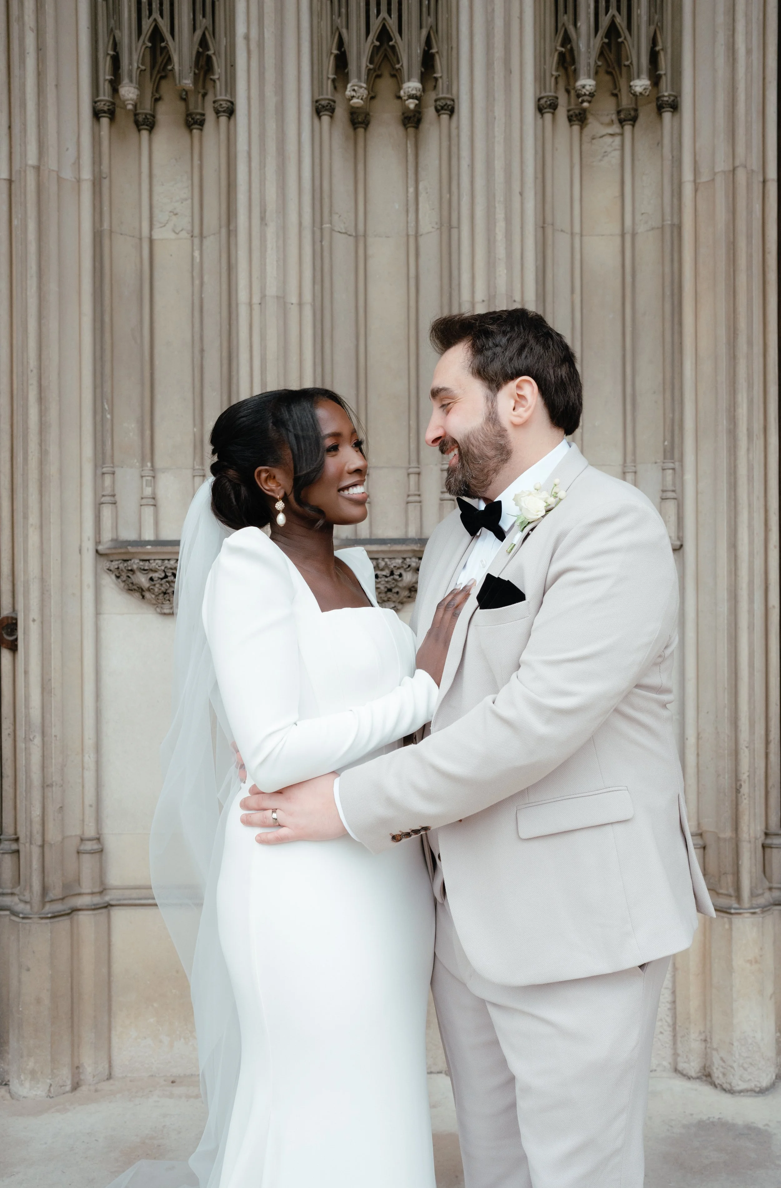 A couple dressed in wedding attire, standing close together outdoors. The woman is wearing a white wedding dress with long sleeves, earrings, and a veil. The man is wearing a light gray suit with a black bow tie and a white boutonniere. He is holding a bouquet of white roses and greenery, smiling at the woman.