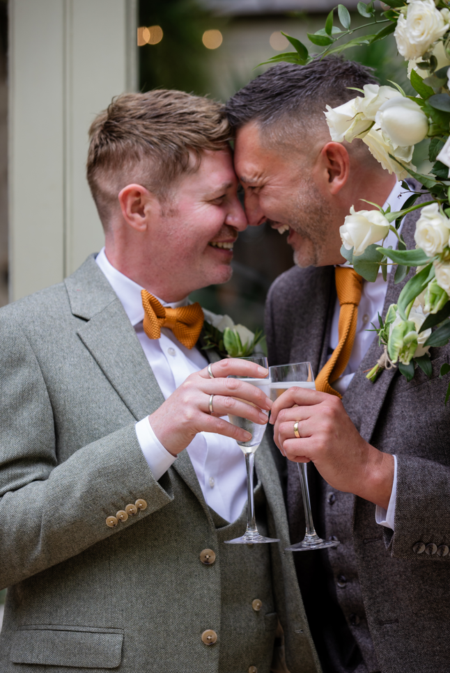 Two men at a wedding, touching foreheads, holding champagne glasses, smiling, dressed in suits with bow ties, surrounded by white flowers and greenery.