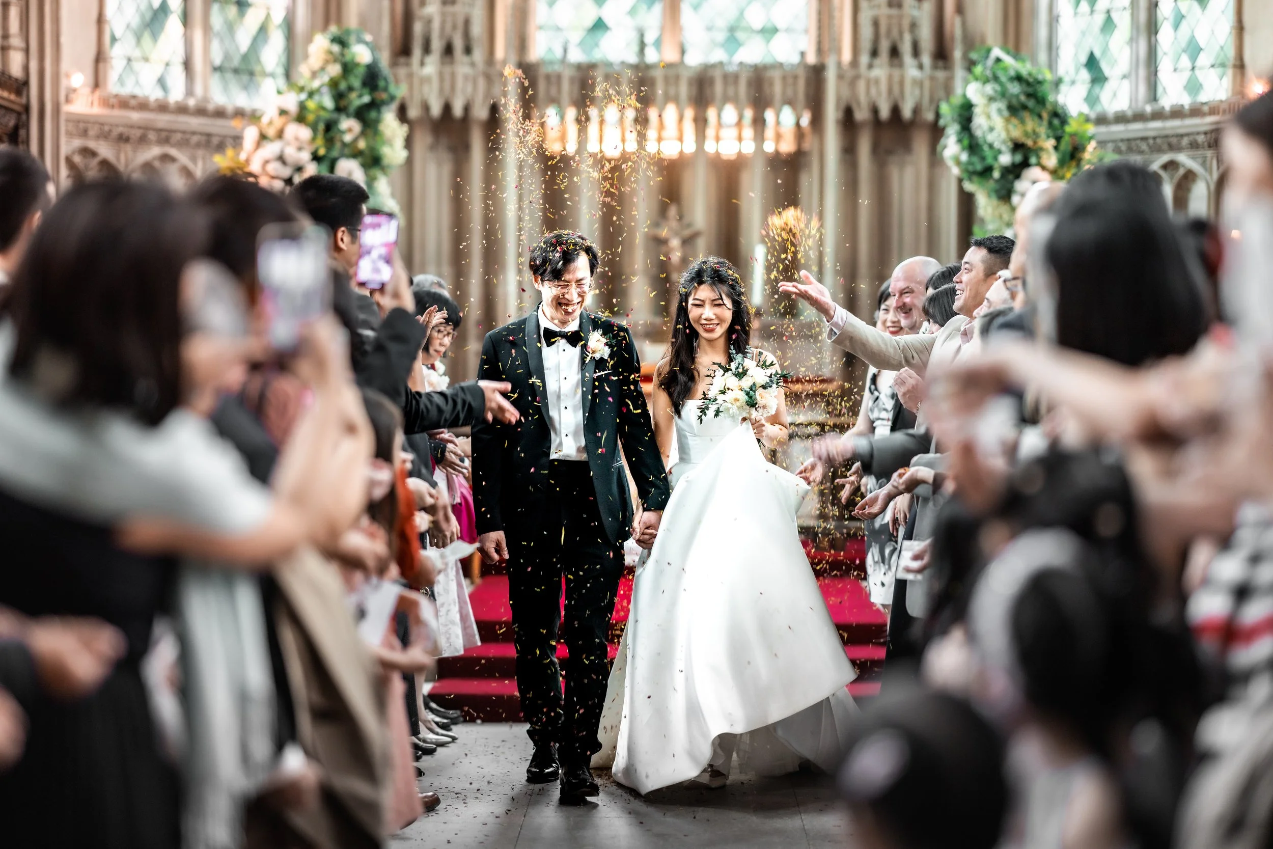 A bride and groom walking down the aisle after a wedding ceremony inside a church, surrounded by wedding guests. Confetti is falling on them and they are smiling.