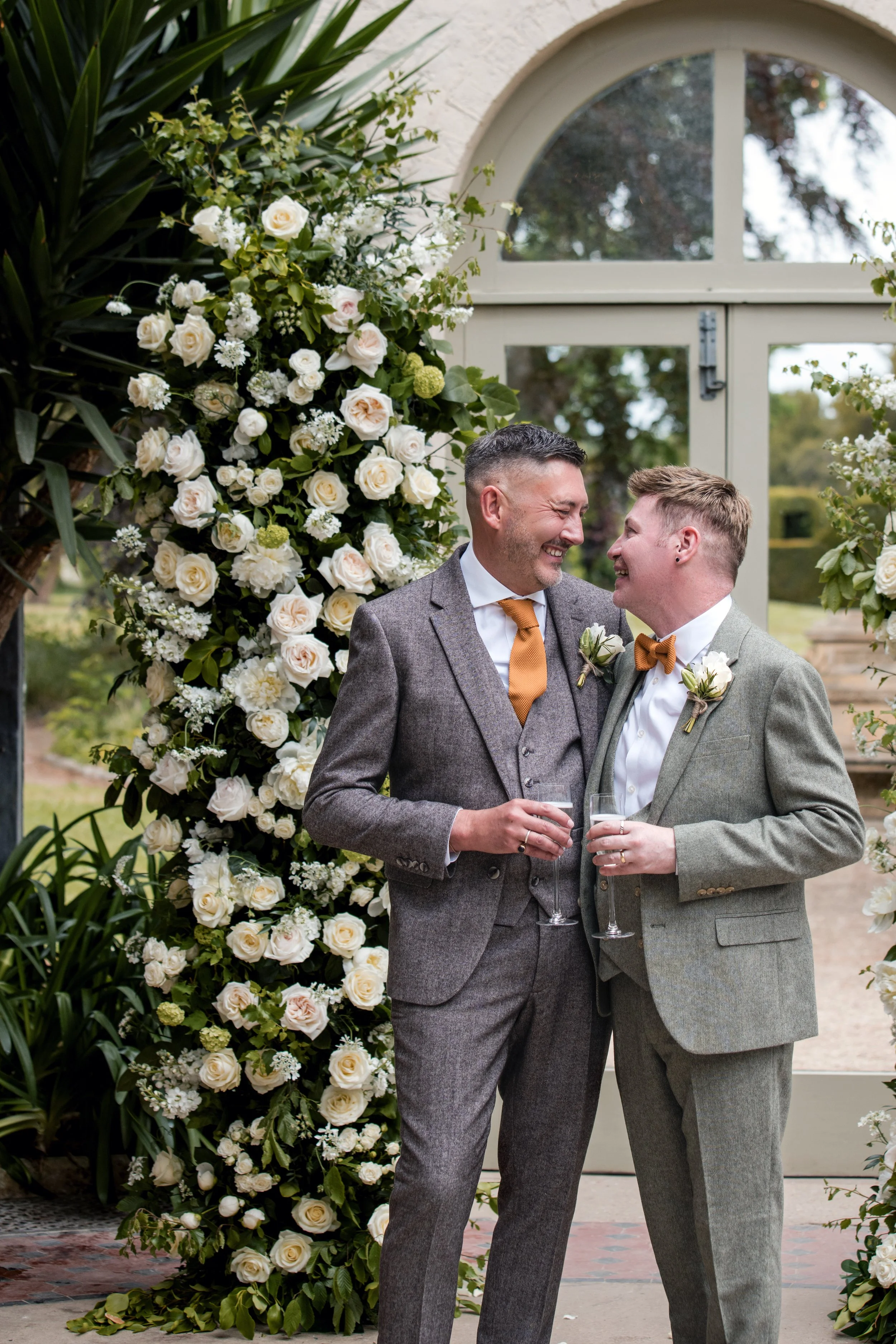 Two men dressed in suits and bow ties, smiling at each other, holding glasses of champagne, standing outside near a floral wedding arch with white roses and greenery, with a window and trees in the background.