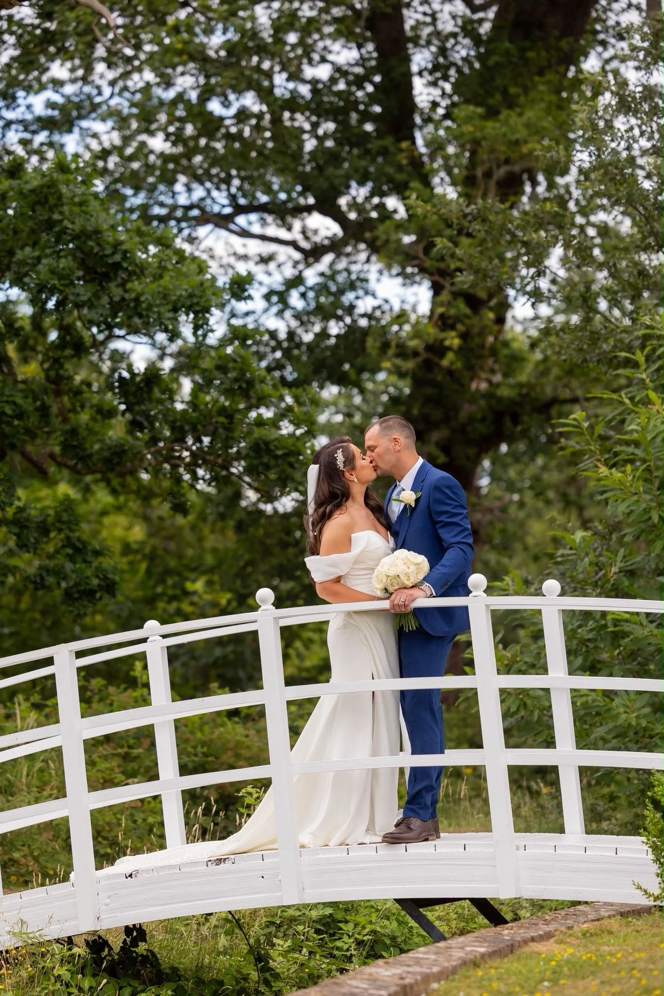 A bride and groom sharing a kiss on a white bridge surrounded by greenery, with large trees in the background.