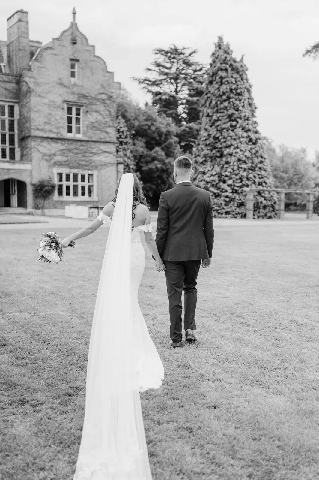 A bride and groom walk hand in hand on a lawn, with the bride holding a bouquet and wearing a long dress and veil, while the groom is in a suit, near a historic building with trees in the background.