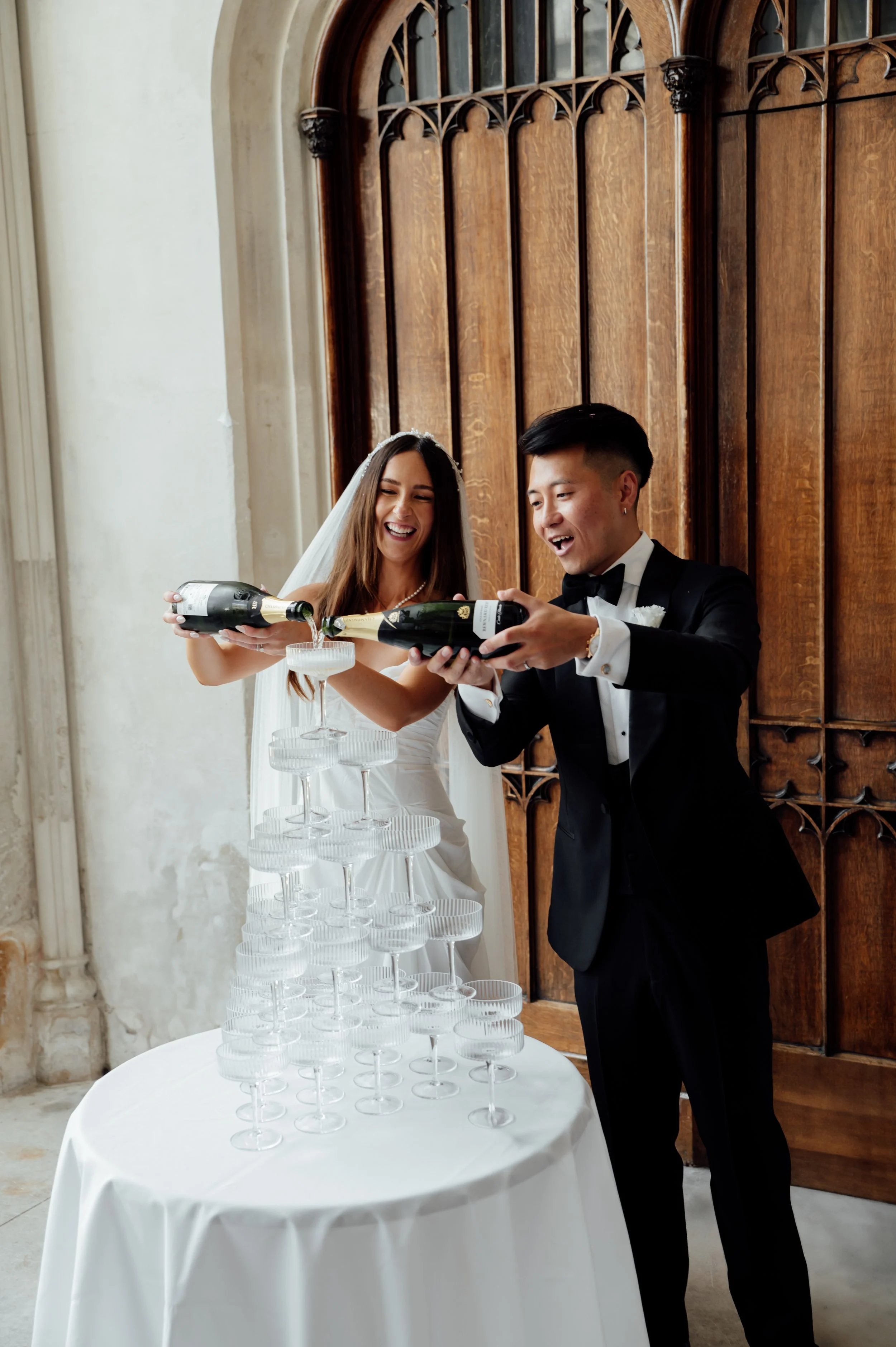 A bride and groom in formal attire pouring champagne into a pyramid of glasses at a wedding reception, standing in front of a wooden decorative wall.