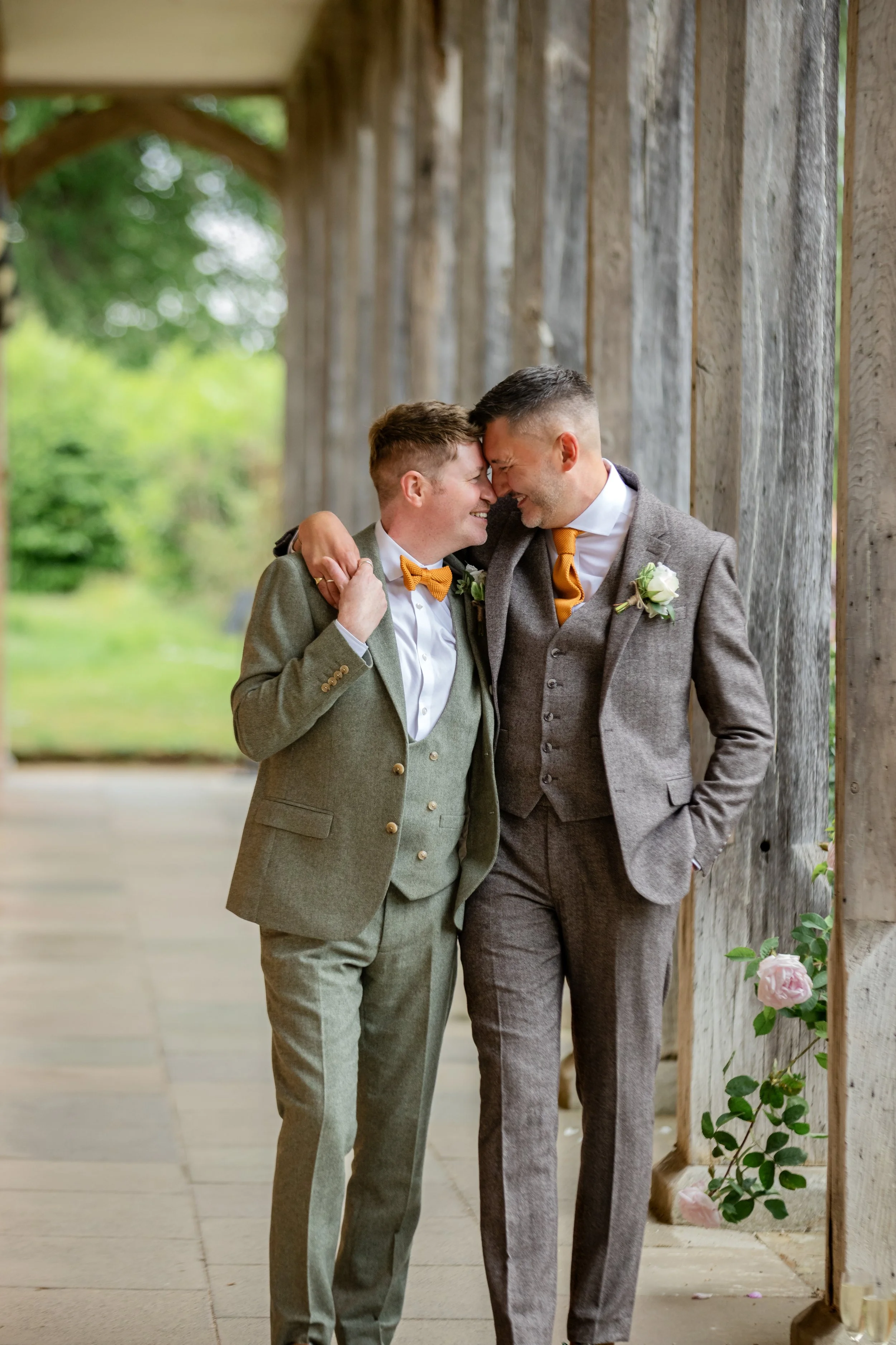 Two men in suits smiling and touching foreheads, standing close together outdoors near a wooden fence decorated with pink roses.