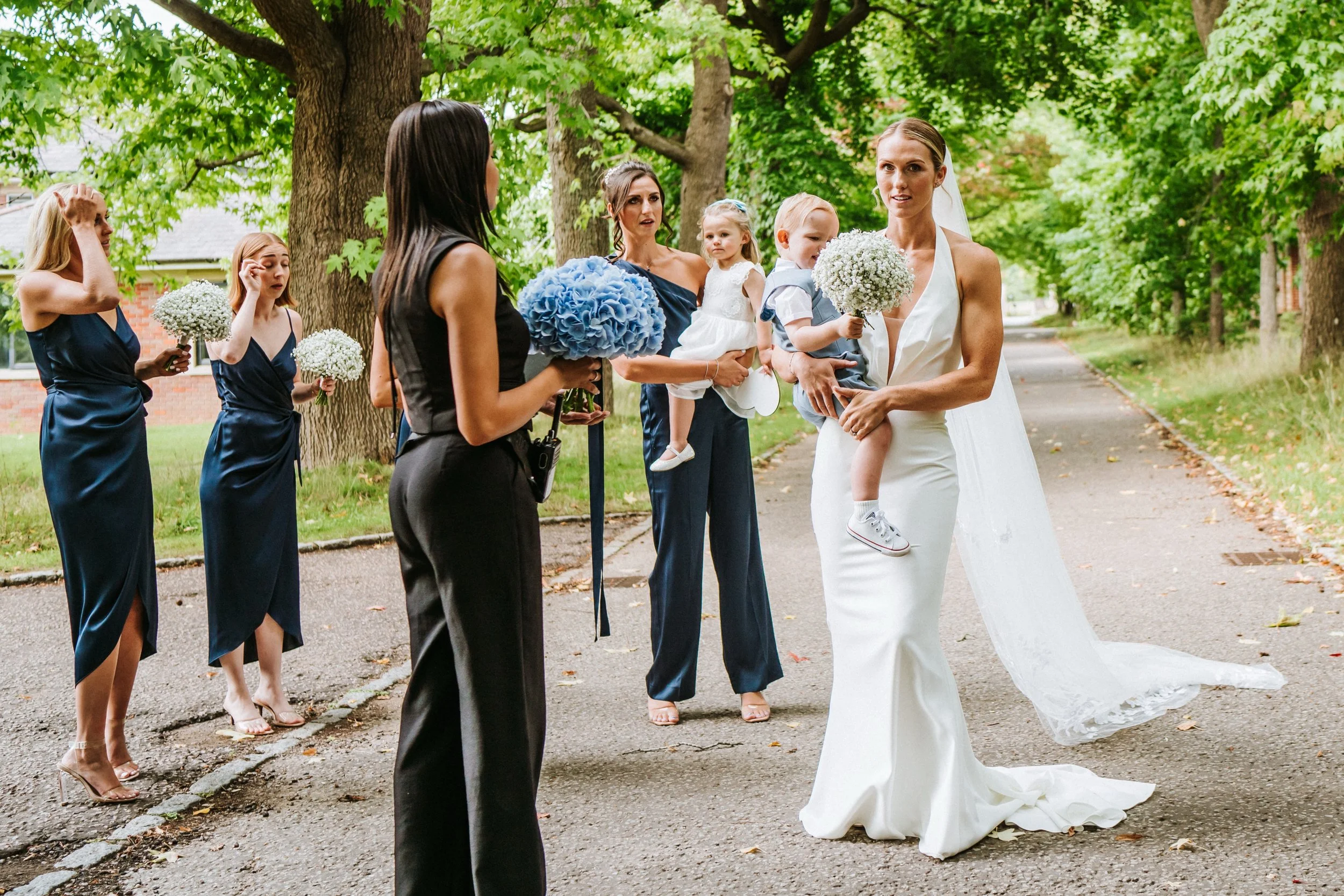 A bride in a white wedding dress holding two small bouquets of white flowers and a young boy with a bouquet, surrounded by women in navy dresses and a woman in black, outdoors near trees and a sidewalk.