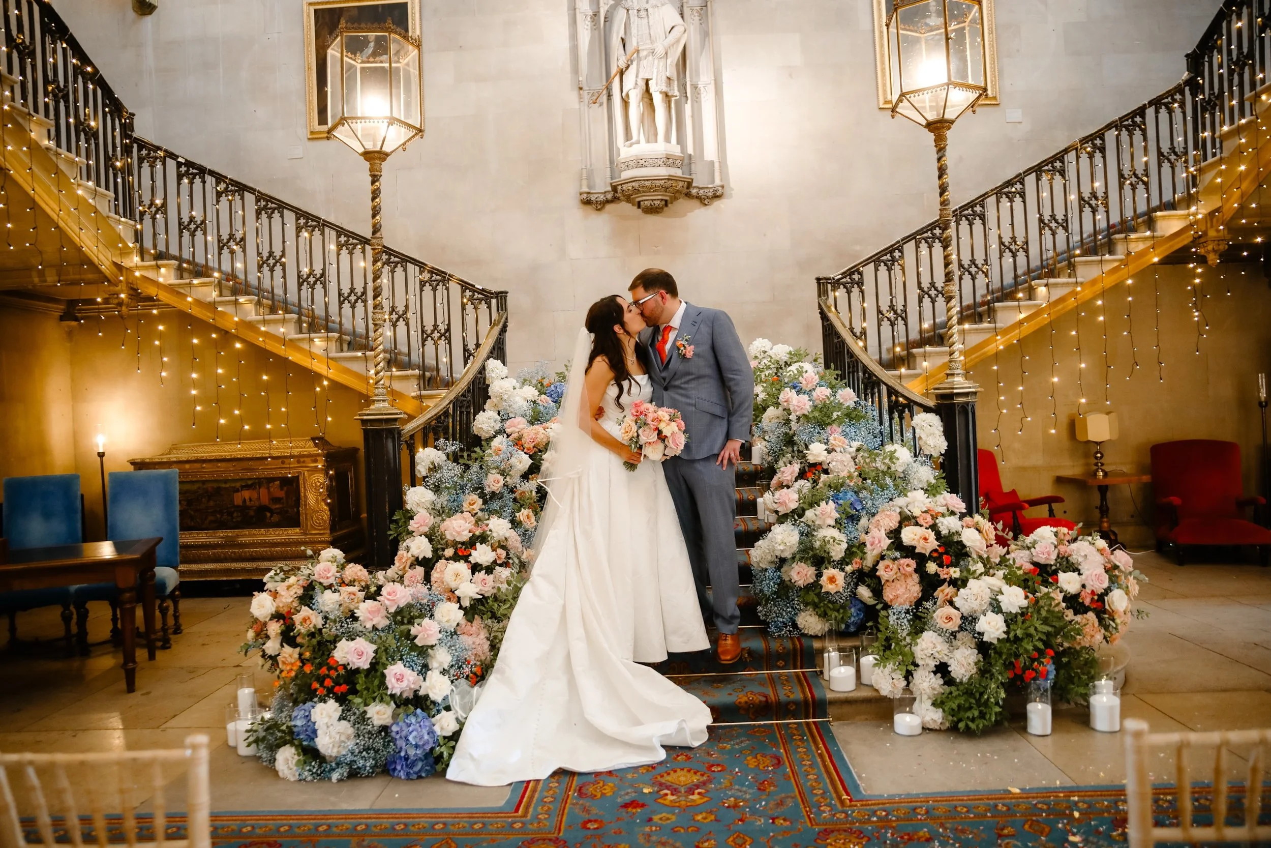 A bride and groom kissing during their wedding ceremony on a floral decorated altar inside a grand hall with staircase and fairy lights.