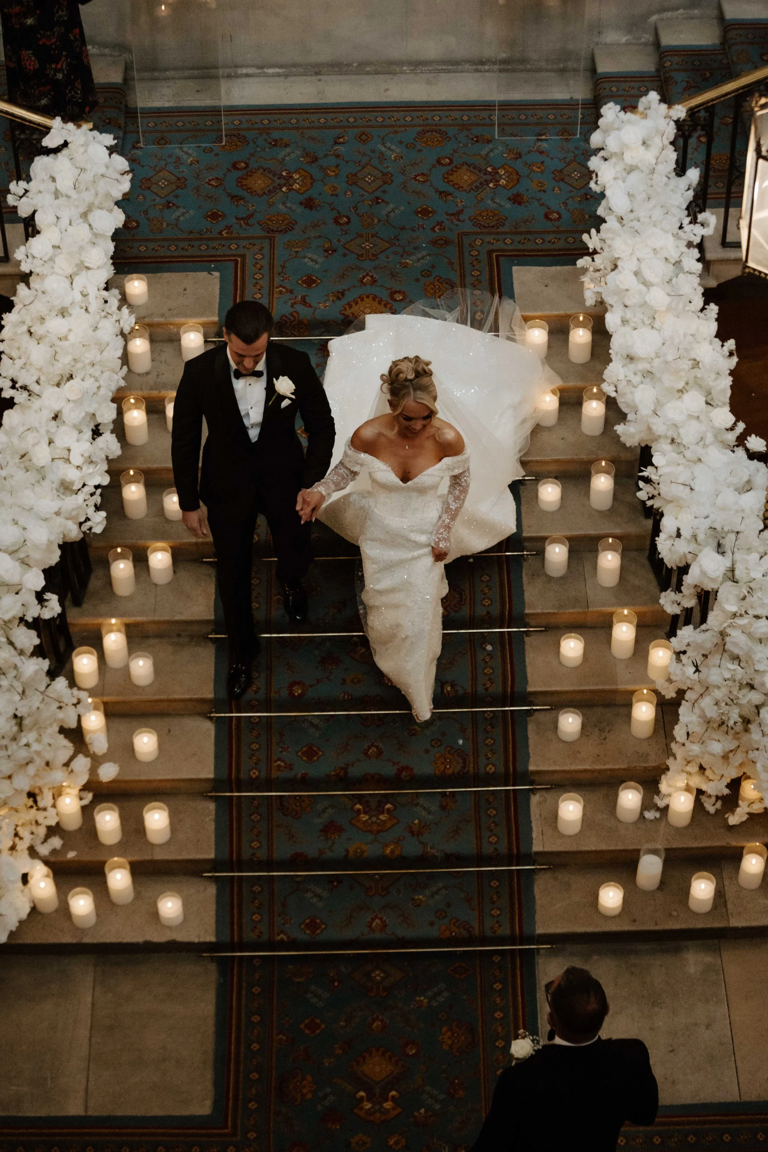 A bride and groom walking down the aisle in a wedding ceremony, surrounded by white floral arrangements and candles on stairs.