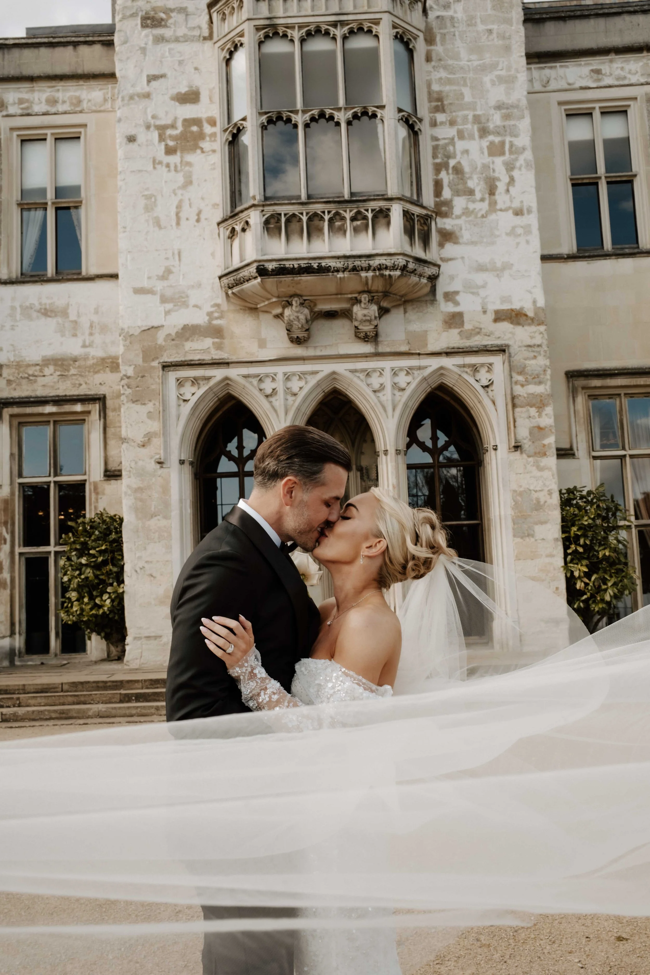 A bride and groom share a kiss in front of a historic stone building with gothic windows and ornate architectural details, during their wedding.