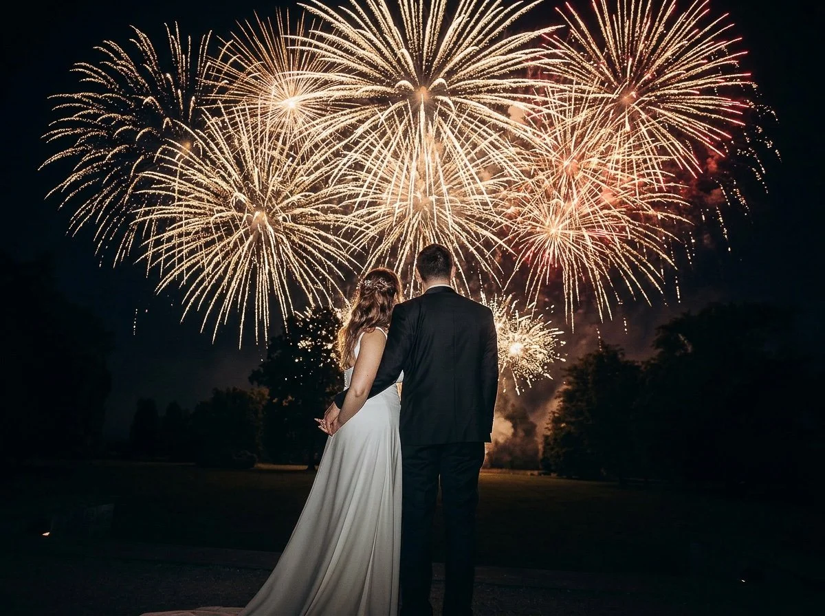 A couple in wedding attire watching fireworks display at night.