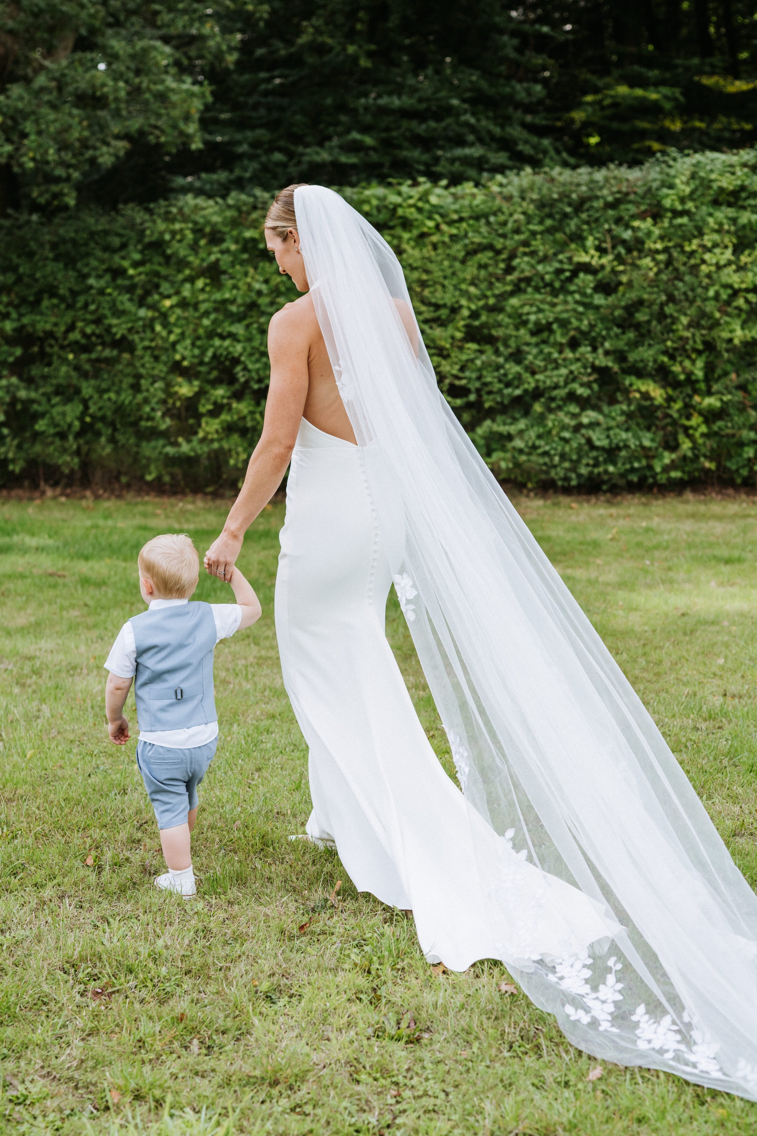 A bride in a white wedding dress with a long veil holding hands with a young boy in a light blue vest and gray shorts in an outdoor setting with green grass and bushes.