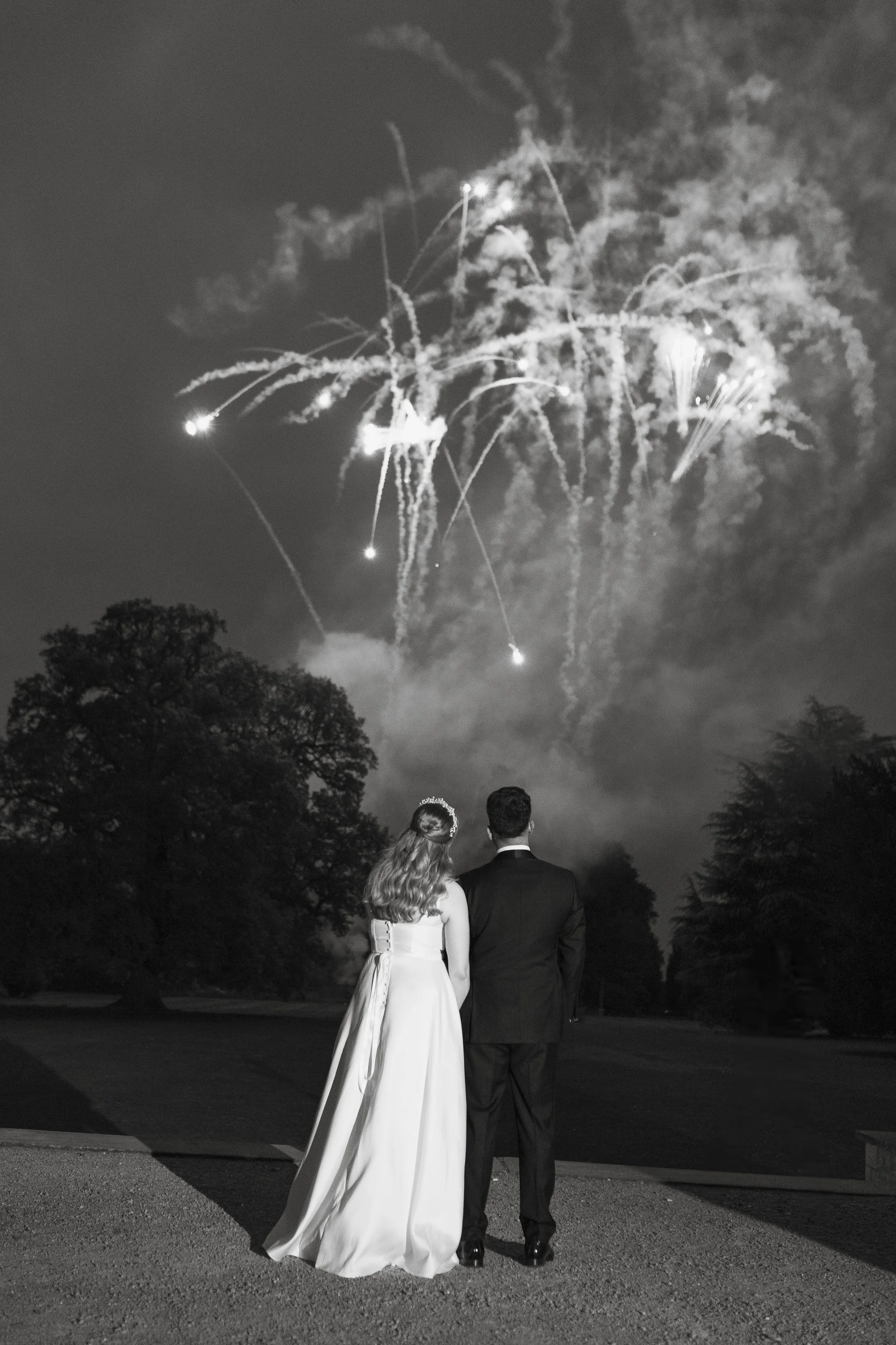 A bride and groom stand together outdoors at night, watching fireworks in the sky. The bride wears a long white gown with a veil, and the groom in a dark suit. Trees are visible in the background.